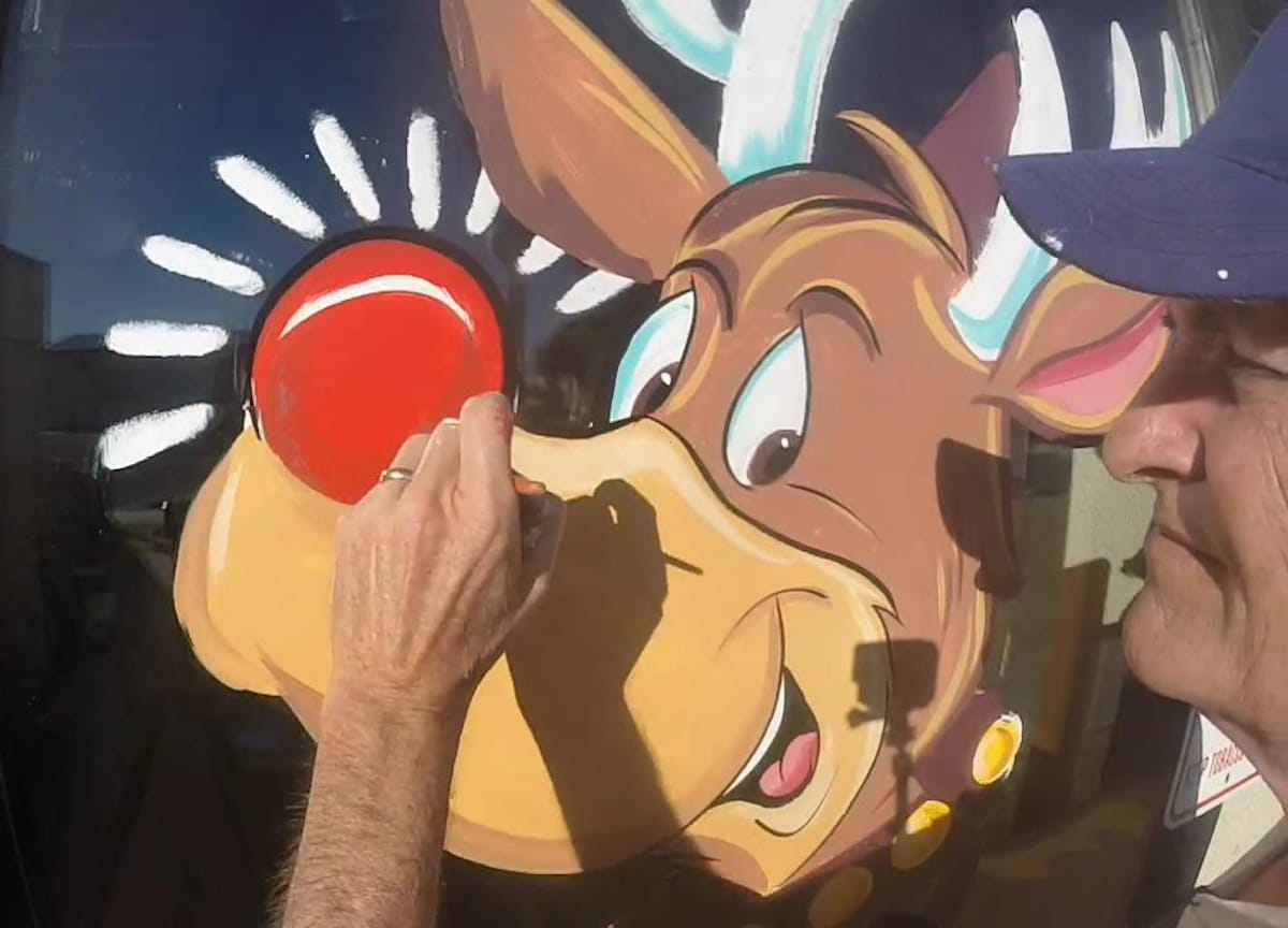 Man painting the red nose on a picture of Rudolph on a window.