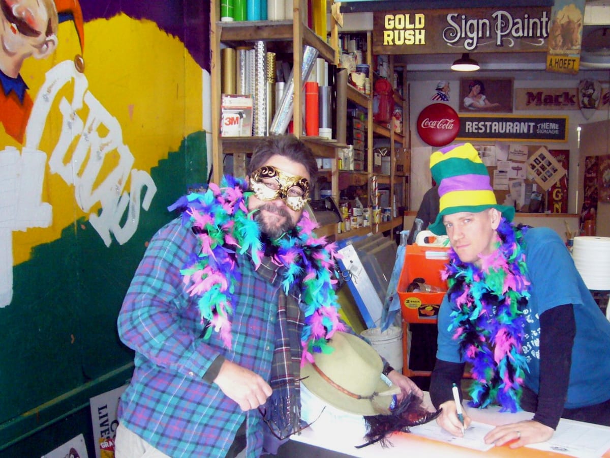Two men with feather boas around their necks, in addition to other Mardi Gras-themed fancy dress. They're posing for the photo in a sign shop adorned with various pieces of lettering and signs on the walls.