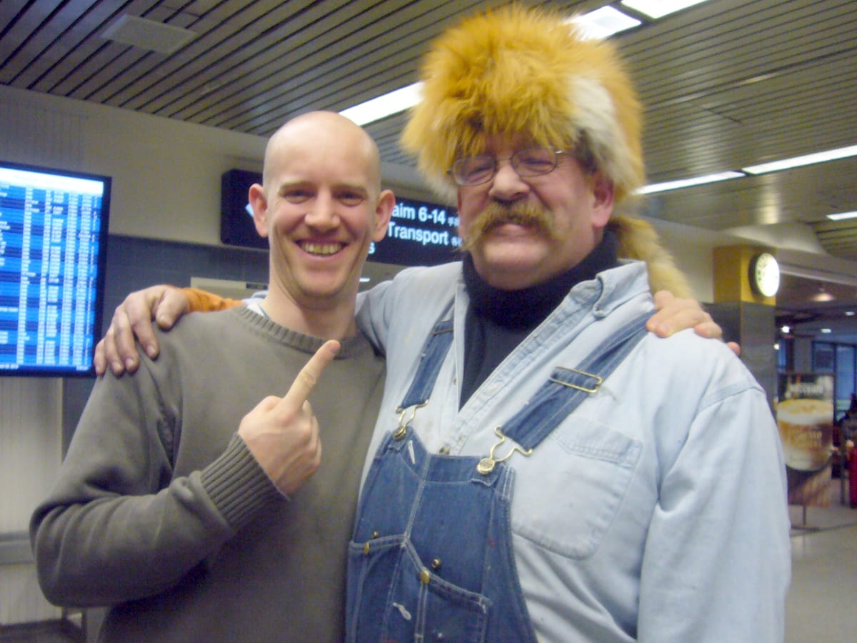 Two men with arms over shoulders in an airport. One is wearing a fox fur on his head while the other points towards it.