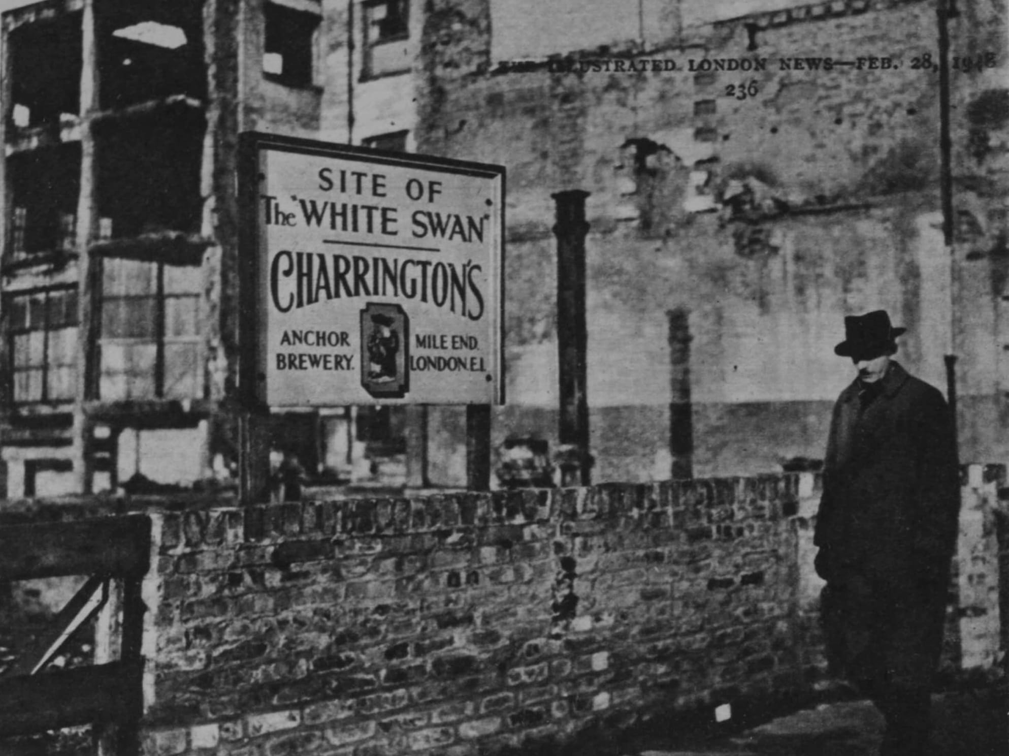 A man in black hat and coat walking in front of a wall with a hand-painted sign protruding above it on two posts. There are destroyed buildings behind the sign, which reads "Site of the 'White Swan'. Charrington's. Anchor Brewery, Mile End, London E1."