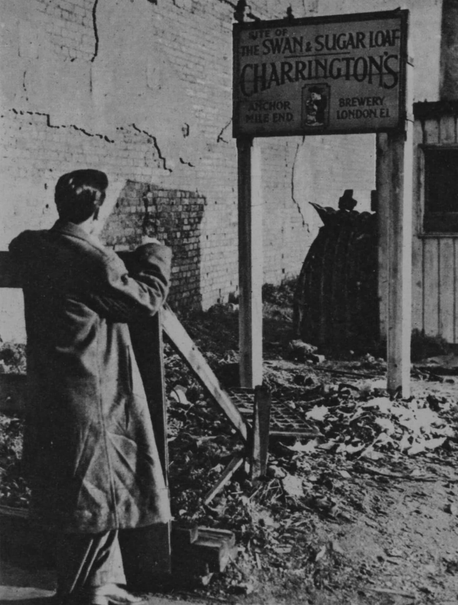 In front of a wall in a poor state of repair, there is an empty plot of land with debris strewn across it. A man is seen from behind looking up at a sign mounted on two posts that reads, "Site of The Swan & Sugar Loaf. Charrington's, Anchor Brewery, Mile End, London E1."