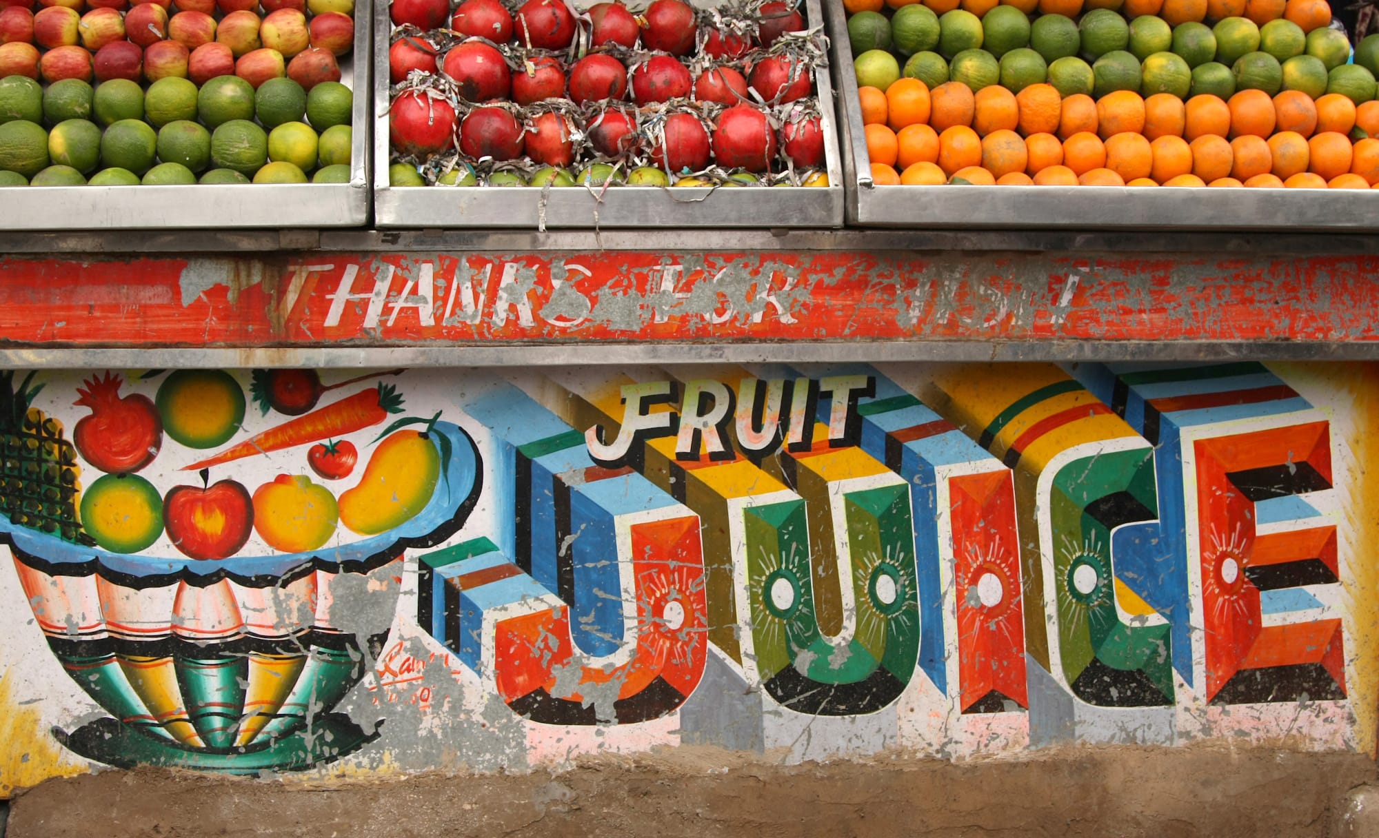 Fruits on display in trays with their colours mirrored below by a flamboyant painted sign that says "fruit juice" next to a picture of a full fruit bowl.