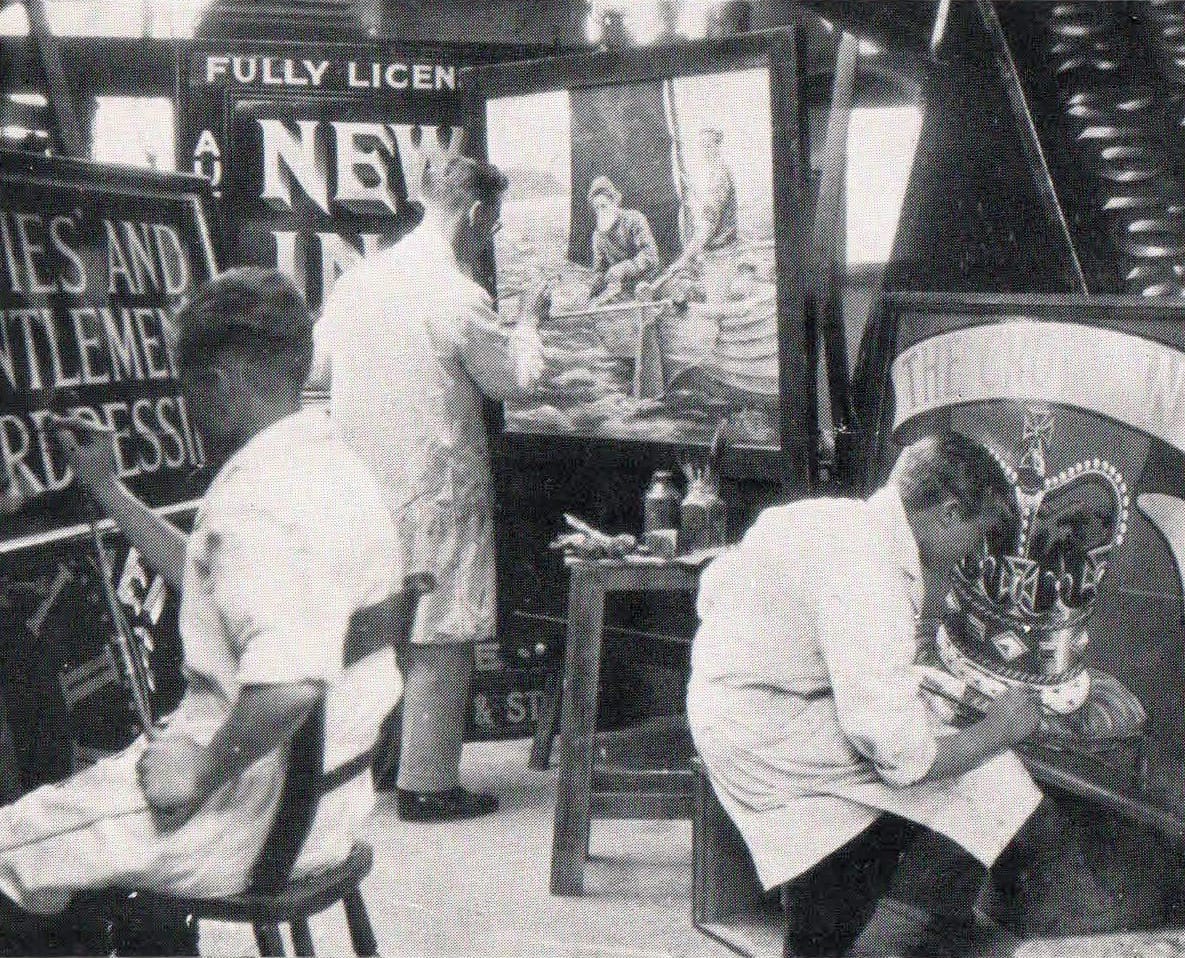 Three people working at easels painting signs with a mixture of lettering and pictorial work. They are quite close together, and the crowdedness of the workshop setting is exemplified by the various pieces of finishing and work-in-progress signage that is propped up all around.