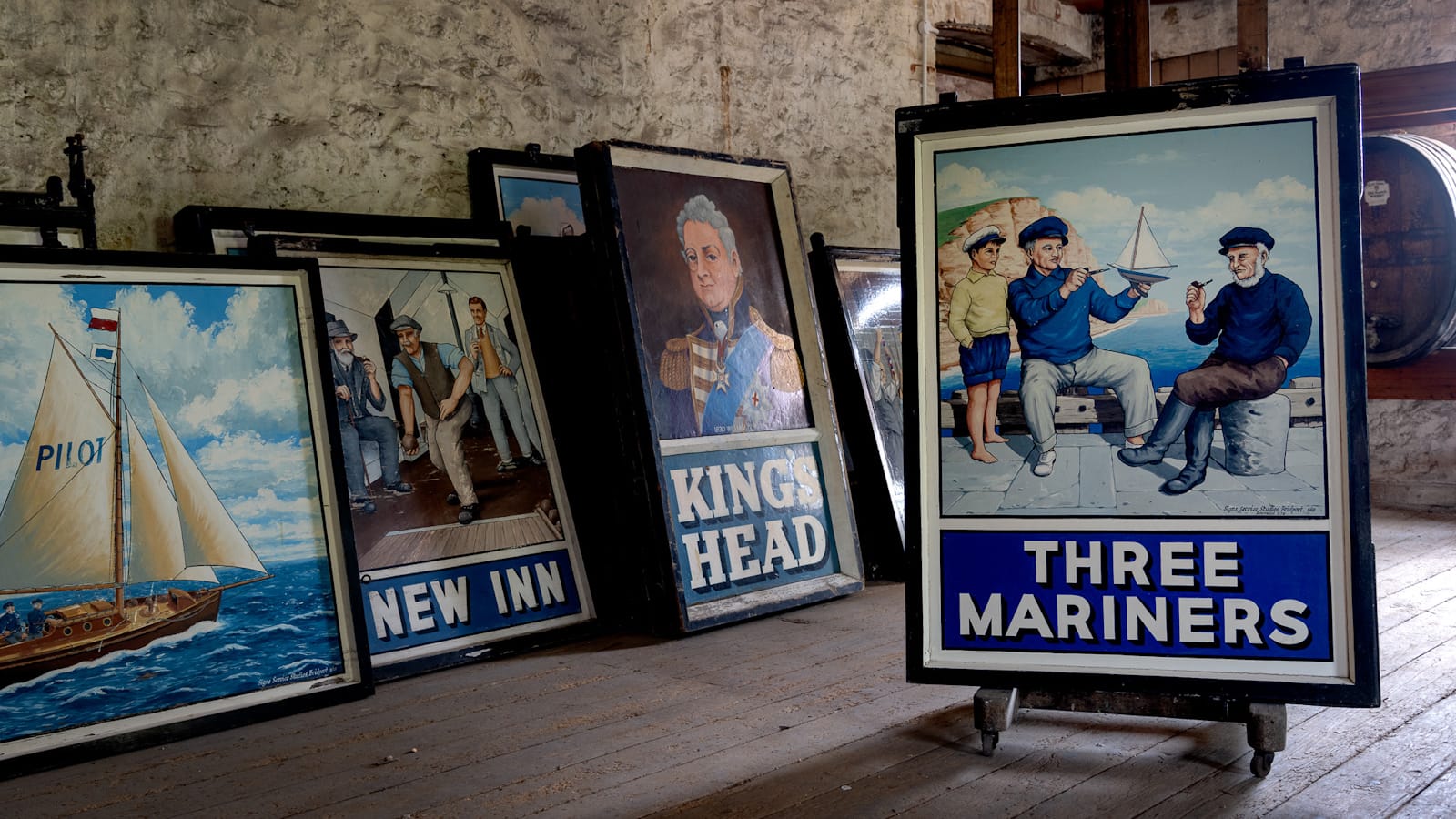 A room with stone walls and wooden floor. Set on an easel in the foreground is a painted pub sign with the bottom third giving the name as "Three Mariners". The upper portion of this sign has a picture of two seated sailors by the sea smoling pipes while showing a model boat to a boy that is standing beside them. Behind this sign, which is propped up on an easel, are various other pub signs sitting against the stone wall, some stacked two or three deep.