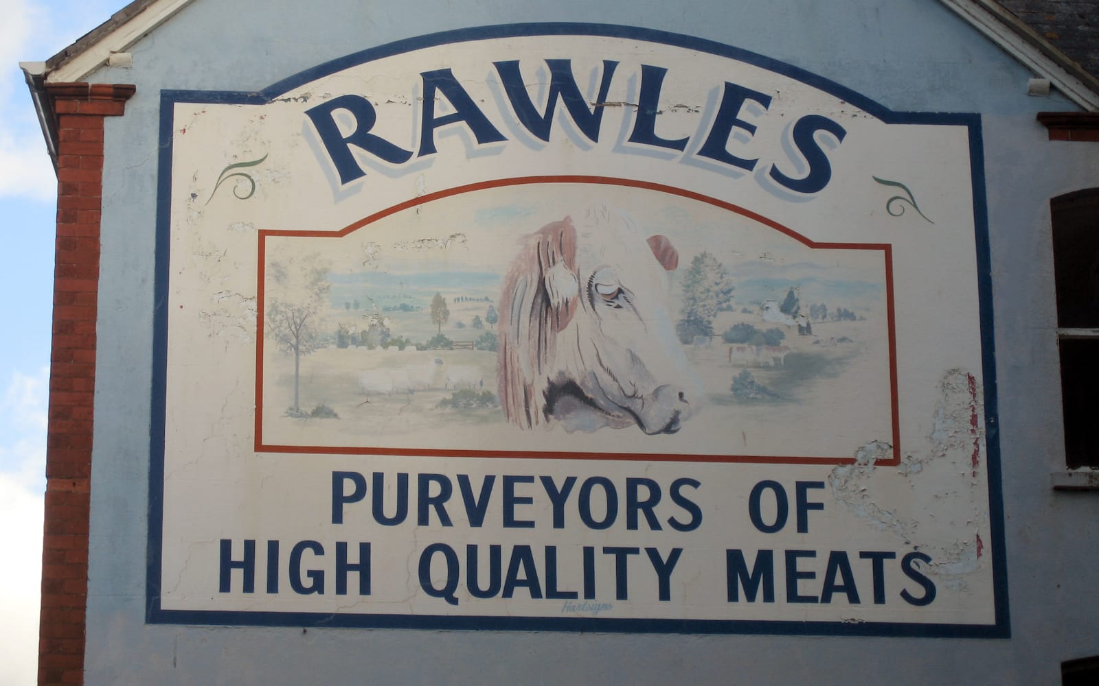 A sign painted on a wall advertising the Rawles butchers. A picture of a cow and a pastoral scene is in the middle, under the name, with lettering in two lines on the bottom that reads "purveyors of high quality meats".
