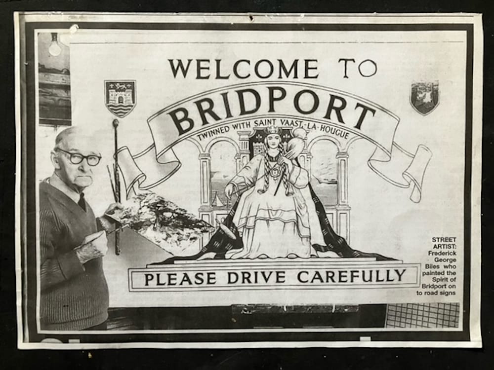Elderly man posing with a painter's palette in front of a 'Welcome to Bridport' sign that he's working on. The sign features a combination of lettering and pictorial elements, which includes the text 'Twinned with Sant Vaast-la-Hougue' and 'Please Drive Carefully'. The picture is of a queen on a throne in a flowing dress and gown, and arched windows behind with sea views. There are also two coats of arms flanking the main lettering at the top.