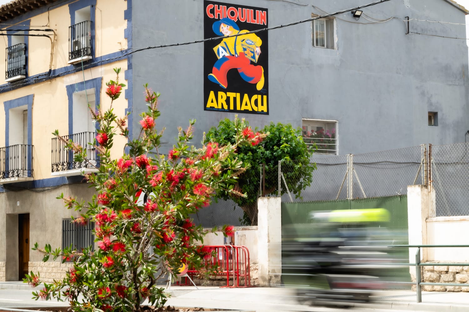 View of a large house with a large side wall painted in grey, but now with with the addition colourful mural of a vintage advertisement for Chiquilin Artiach with a pictorial of a book running with two boxes of biscuits under his arms.