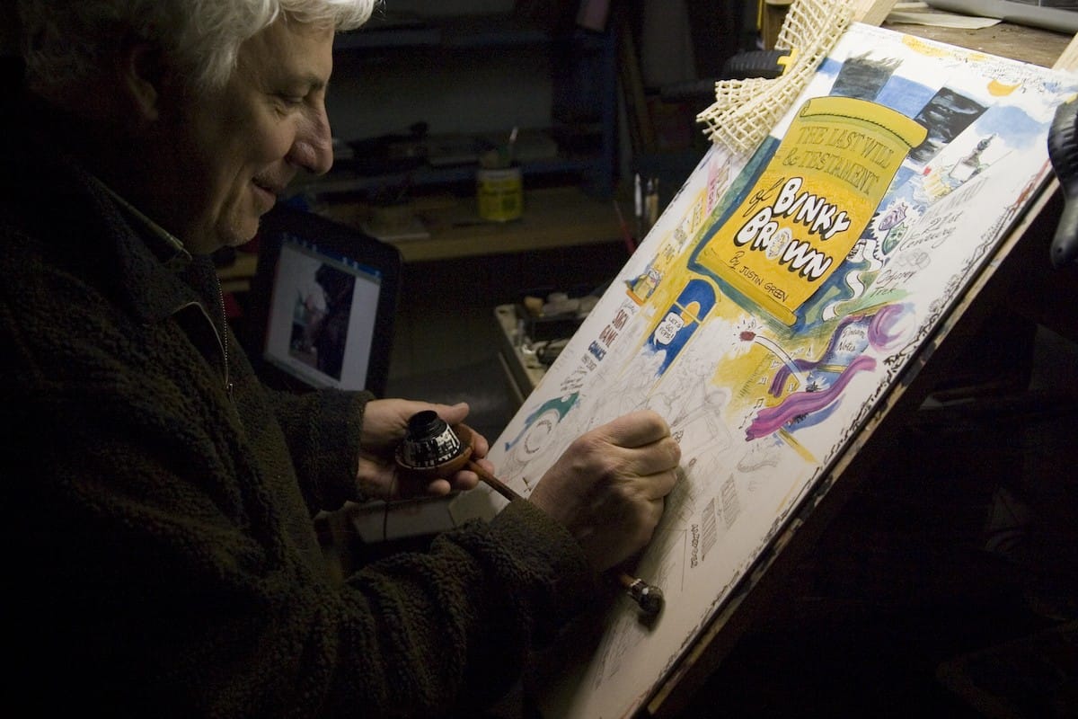 Man at an easel painting. It can be seen that the piece he's working on is a comic and the words 'Binky Brown' are visible in bouncy white lettering.