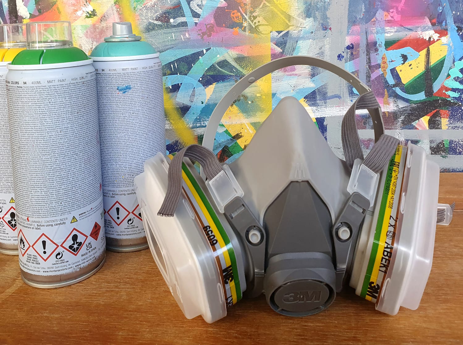 Protective mask with various filters in place, photographed beside three cans of spraypaint and in front of a wall painted with many lines and blocks of sprayed paint.