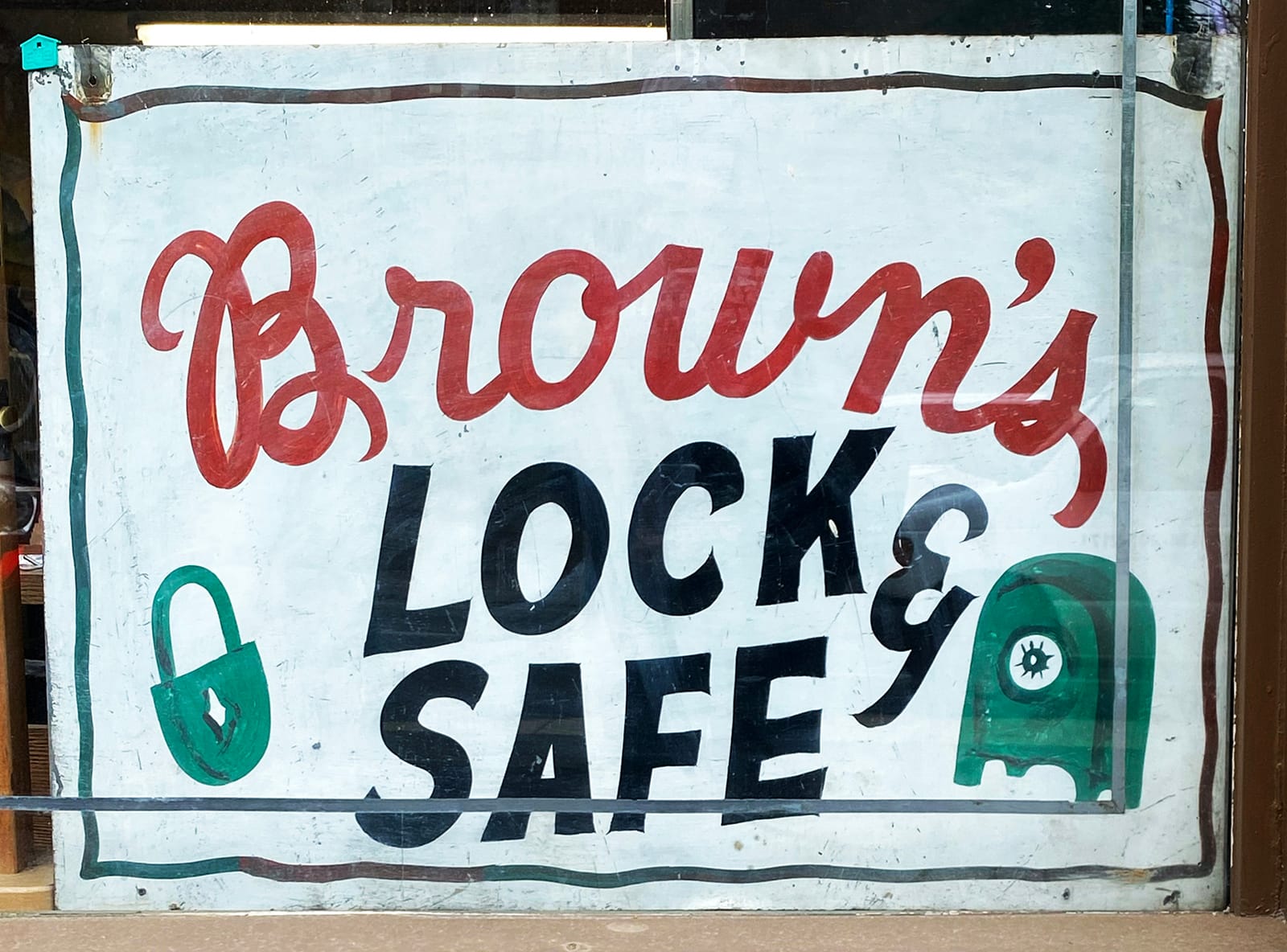 A sign board in a window that is crudely painted in red cursive ('Brown's') and black san-serif ('Lock & Safe') letters. There is a wiggly border placed around these, and small pictorials of a padlock and a safe flanking the 'Lock & Safe' lettering.