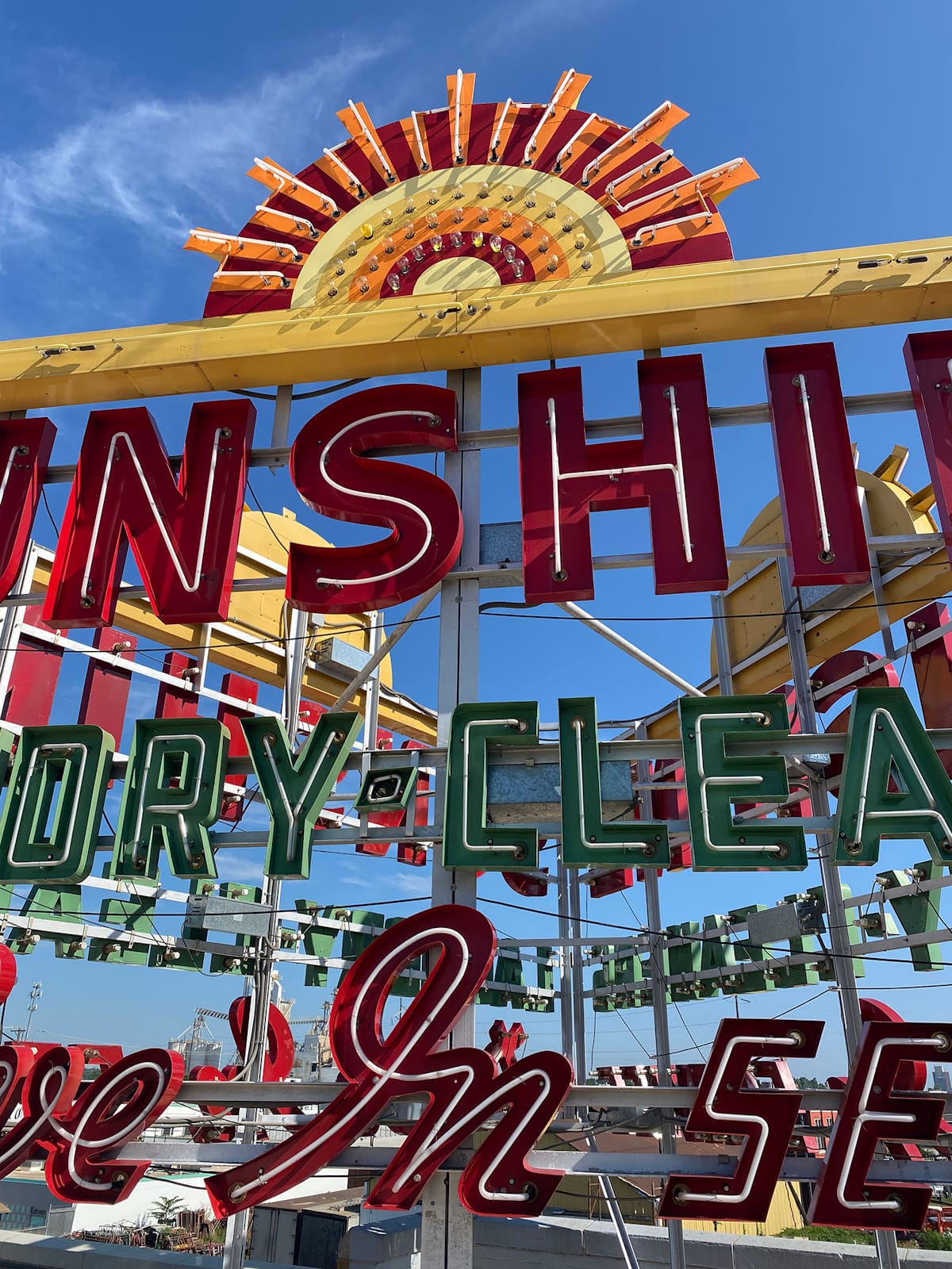 Portion of a neon sign set within channel letters and mounted onto a metal framwork on the top of a building. Looked at from above, the frame would form a triangle, with the same lettering reproduced on all three sides for visibility from all angles. From what's visible, it reads "Sunshire Dry-Cleaners" and what appears to be "Drive-In Service".