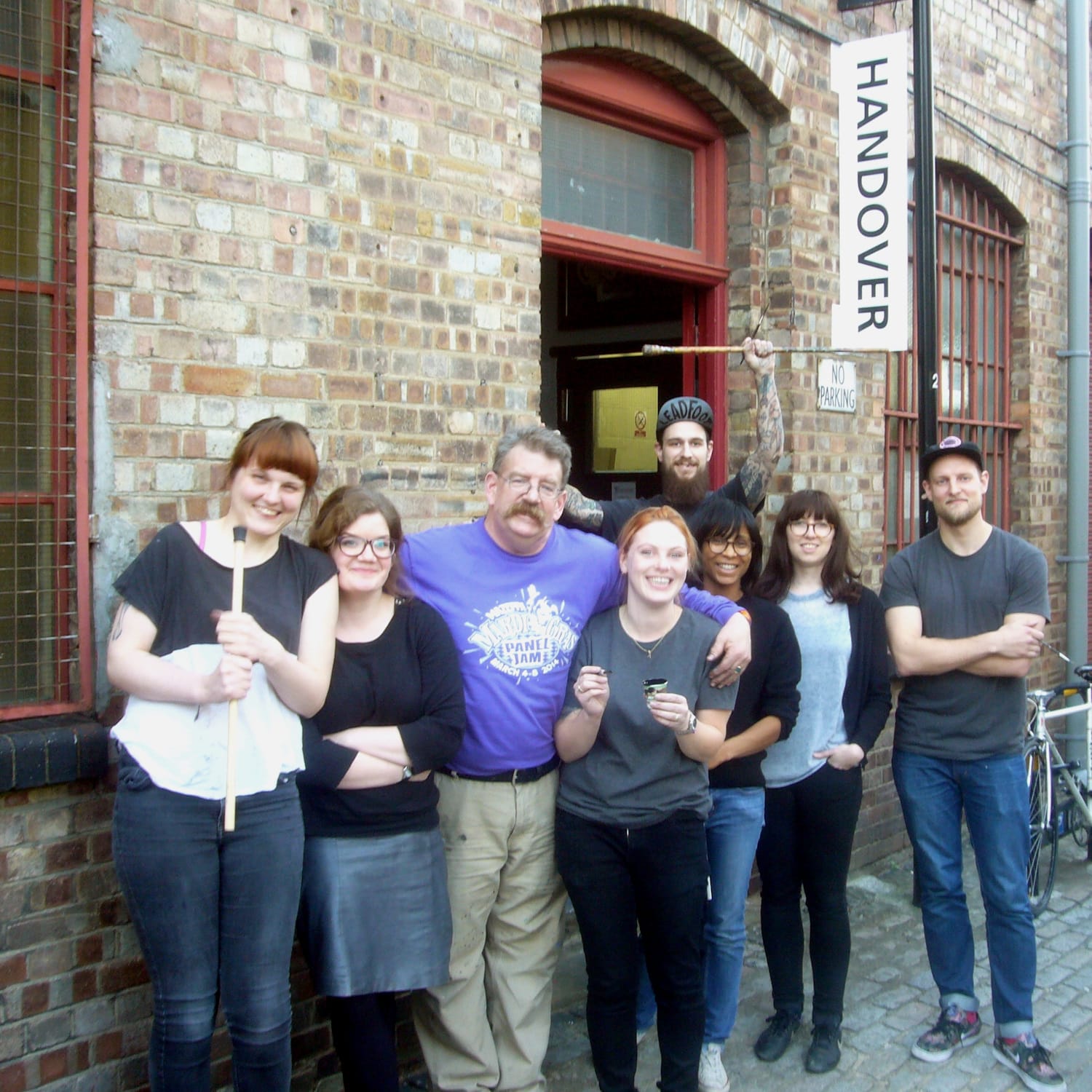 Photo of eight people posing outside a brick building with a protruding sign that says "Handover".