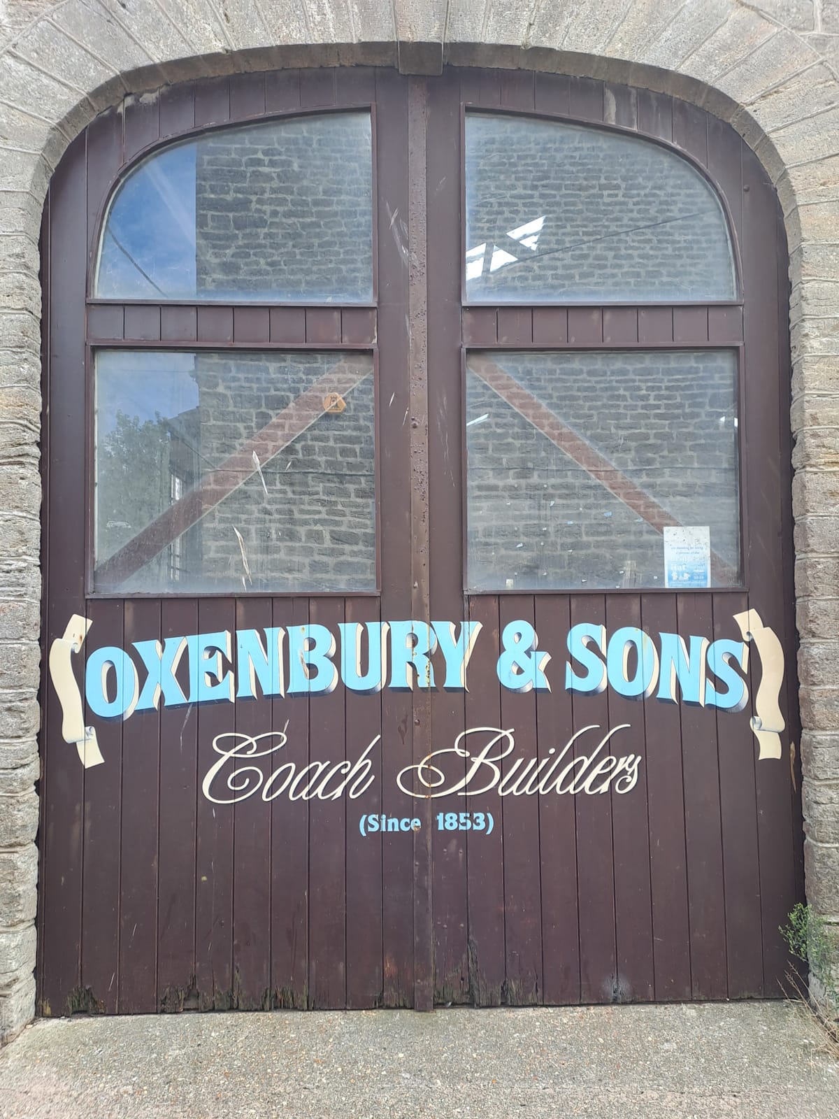 Large brown double doors on a stone building with windows set in the upper portions. On the wooden slats beneath are painted letters advertising "Oxenbury & Sons, Coach Builders (Since 1853)".