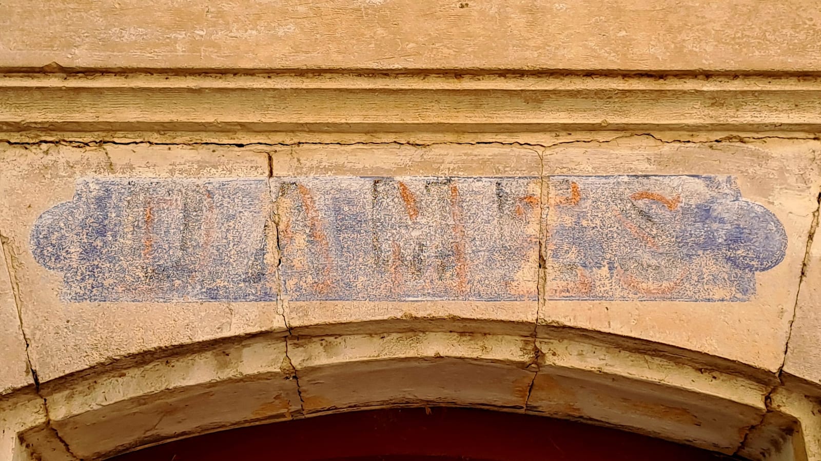 Close-up of stonework above an arched doorway. Set across the keystone, and those either side of it, is a fading panel of paint with lettering in red on a blue background that is legible enough to make out the word 'dames'.