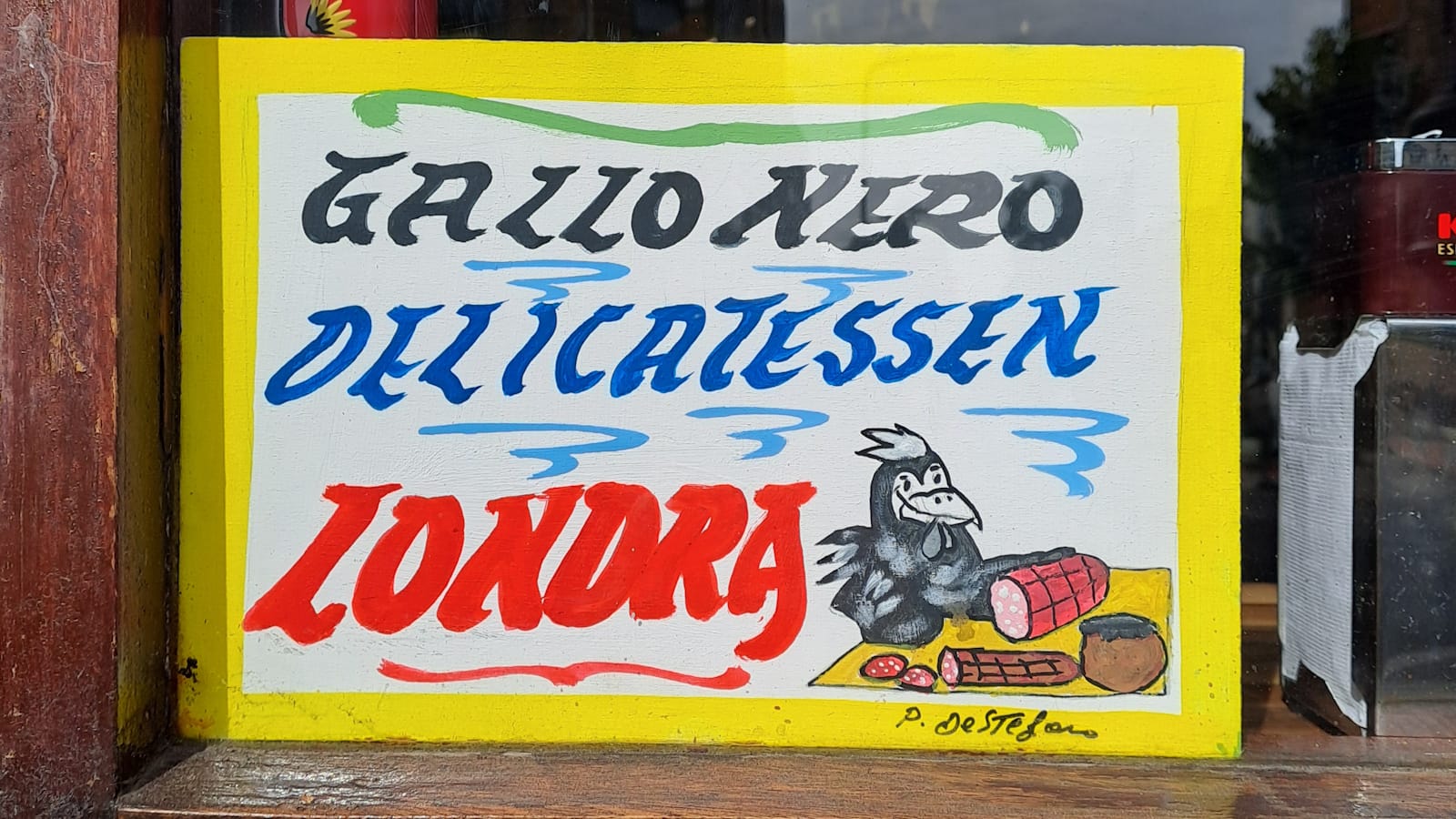 Small hand-painted sign in a window with a yellow border, a pictorial of a chicken cutting ham, and lettering that reads 'Gallo Nero, Delicatessen, Londra' in black, blue, and red lettering respectively..