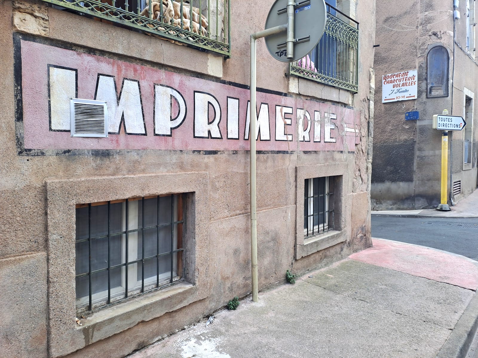 View of stone building at street level. A large fading painted sign runs horizontally between low-level windows and those on the first floor, reading "Imprimerie". It is painted in white letters with a black outline, set on a red background with a black border. Across a small road just beyond the end of the sign is another that has some red lettering for various butchering services, and the business name in a cursive lettering style.