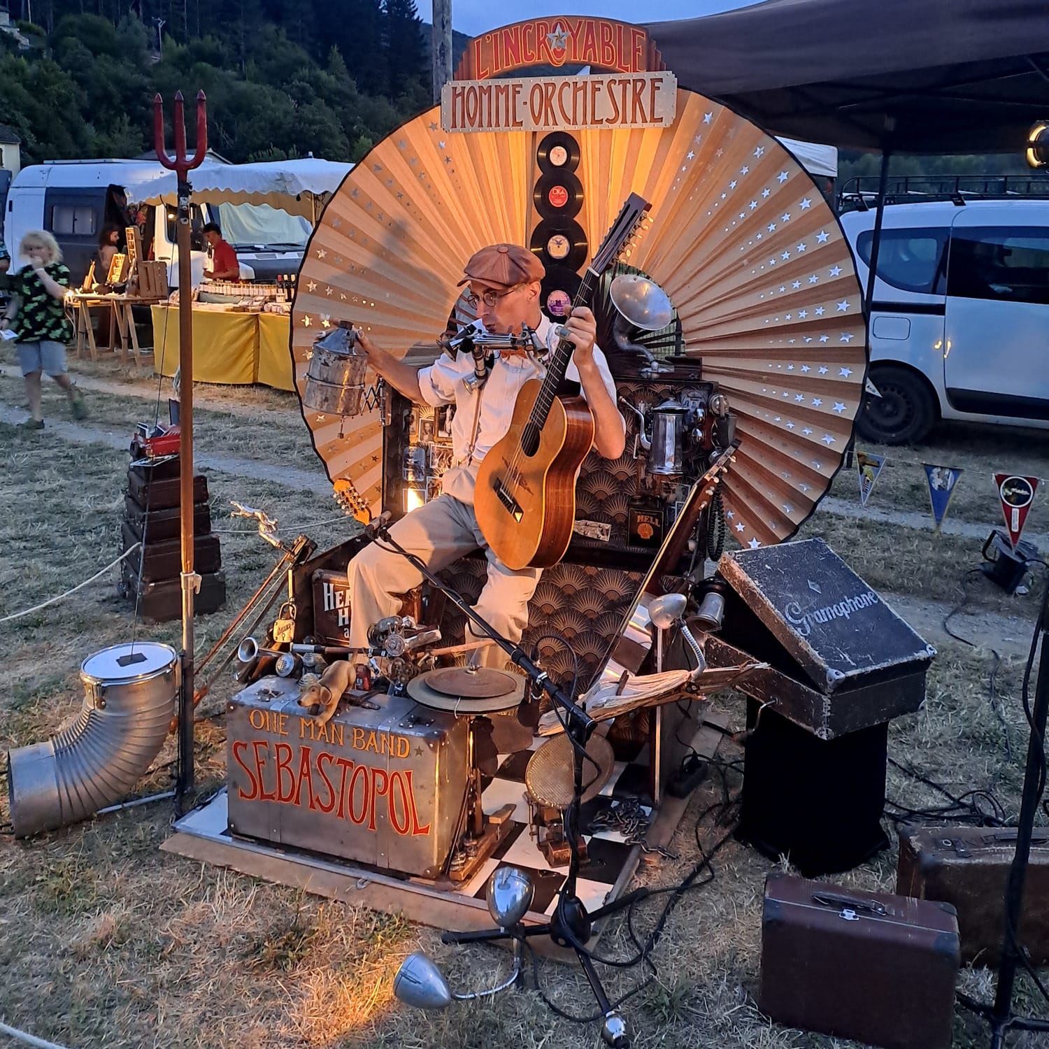 A man holding a guitar and surrounded by an array of different musical instruments and their cases. Behind him is a large circular fan-like structure with a vertical line of vinyl records mounted in the middle and painted signs above that read "L'incroyable homme orchestra". One of the cases immediately in front of his legs is made of metal and painted with "One Man Band Sabastopol".