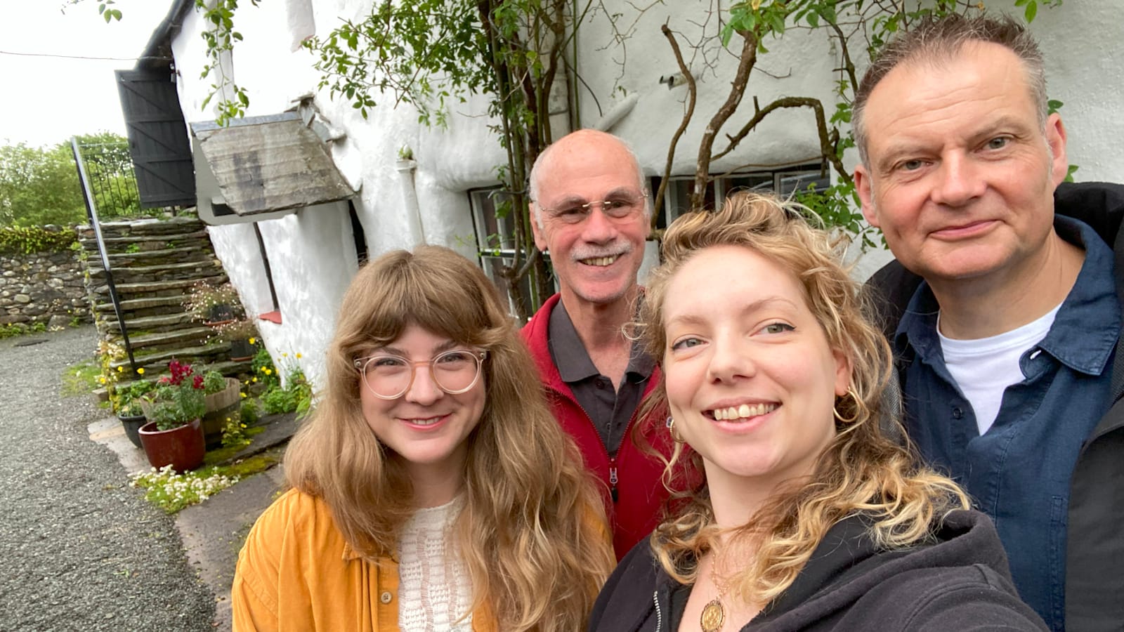 Group of four people smiling for a selfit outside a white-walled building with various plants growing on/beside it.