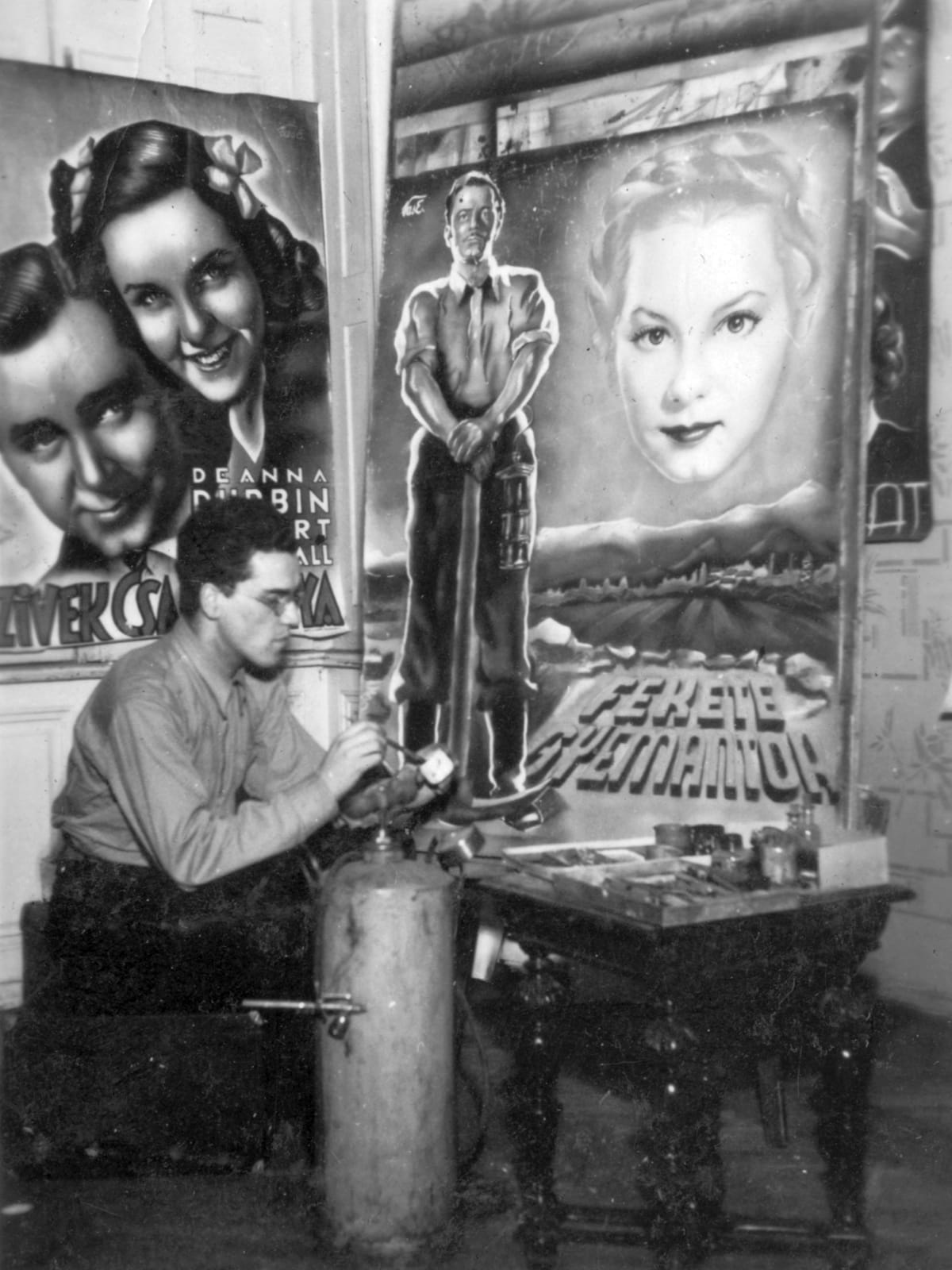 Black and white photo of a man paletting a brush on a paint cup. Beside him is a table with painting materials and a gas canaster with airbrush. Behind him are two painted film posters.