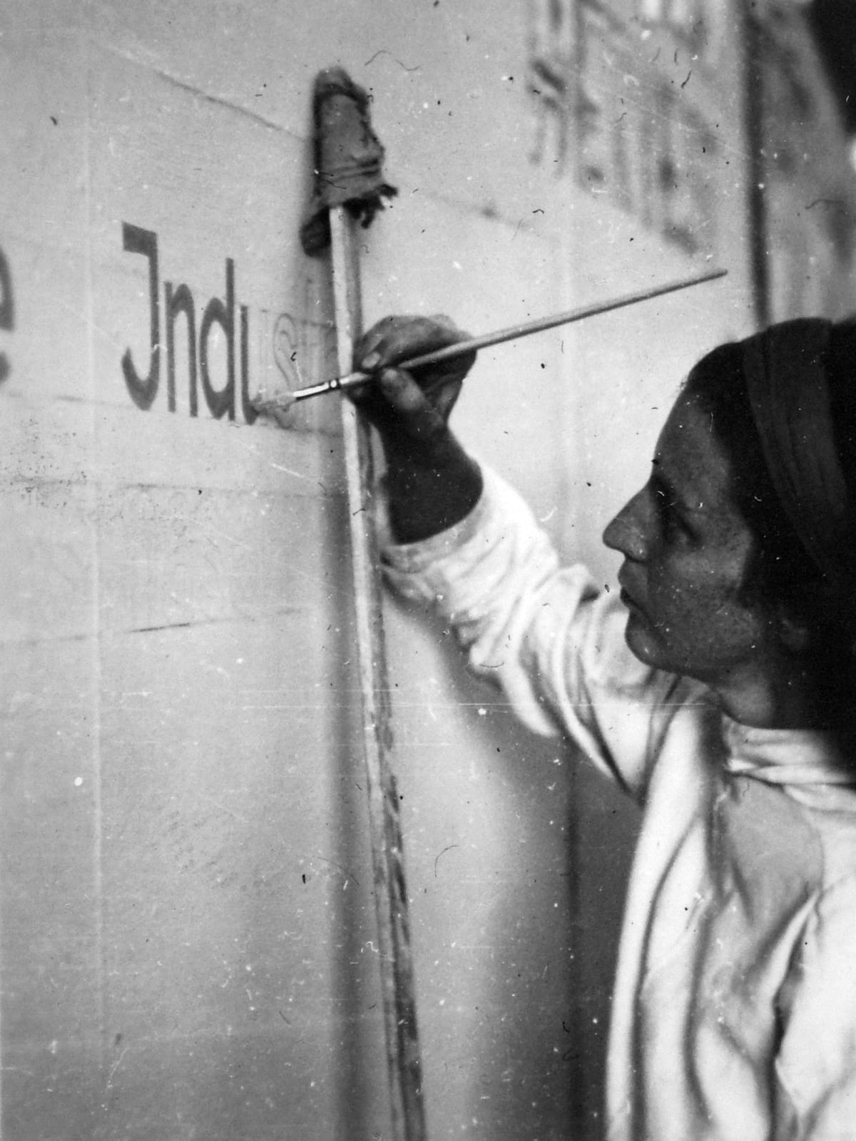 Grainy black and white photo of a women resting her hand on a large, vertically positioned mahl stick while she paints letters that are outline on the wall.