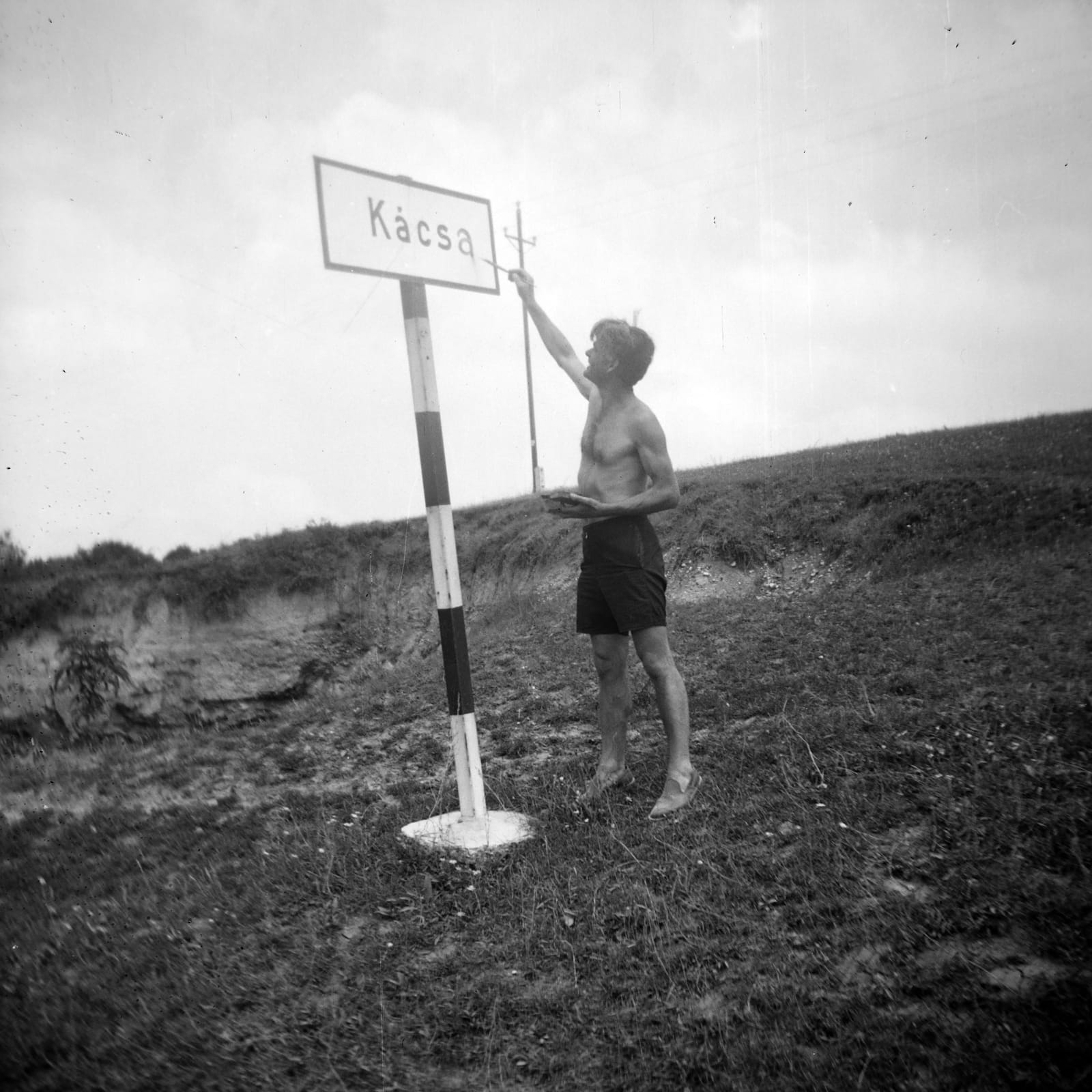 Black and white photo of a man wearing just black shorts reaching up to paint an extra letter on a sign with the village name, Kács, to make it read Kácsa.