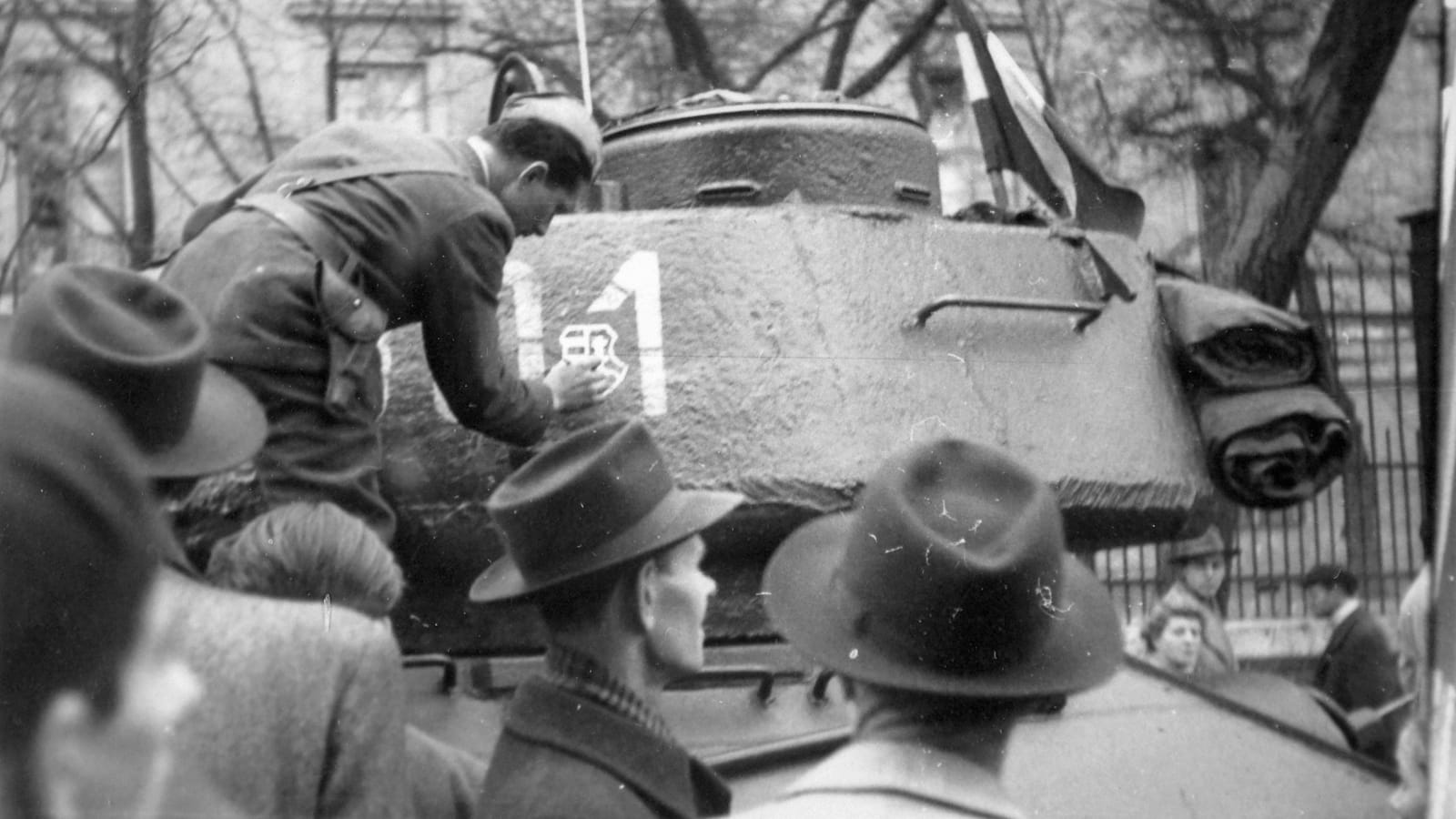 Black and white photo of a man in military uniform painting a crest on a tank that already has numbers painted on it.