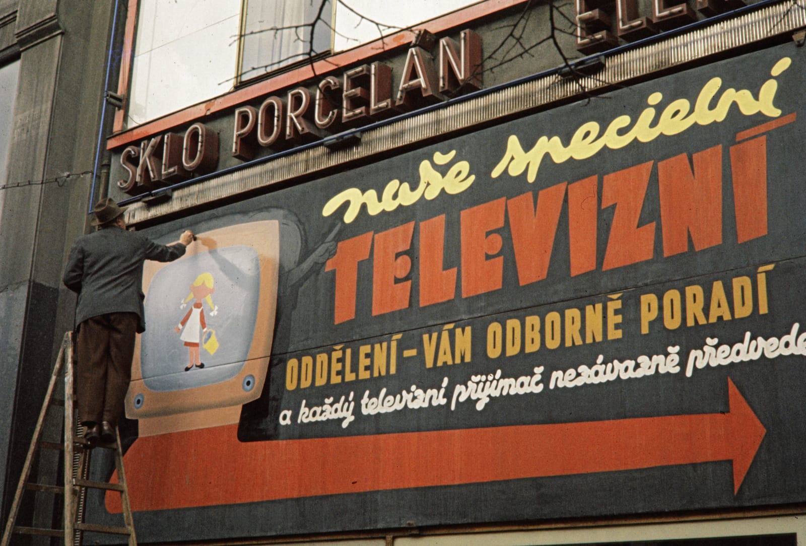 Man on a ladder painting a pictorial of a television for a large billboard advertising televisions. Juat above this are neon letters which seem to be the name of the shop where these televisions are available to buy.