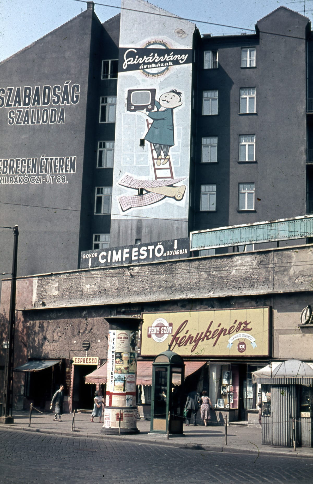 Colour photo of a street scene with shops at street level and then a block of flats behind that are adorned with painted signs on the wall, at least one of which is advertising televisions.