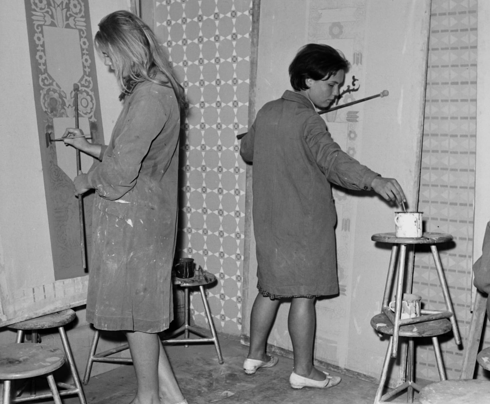 Black and white photo of two women in painters coats working with mahl sticks and brushes on heraldic panels.