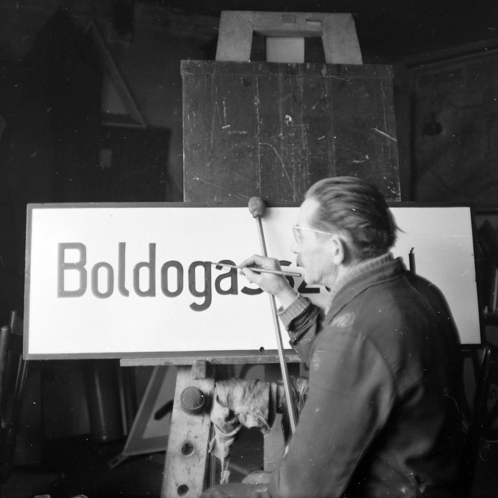 Black and white photo showing the view from behind a man sat at an easel painting black lettering on a white sign board with mahl stick and brush. The lettering appears to be a place name.