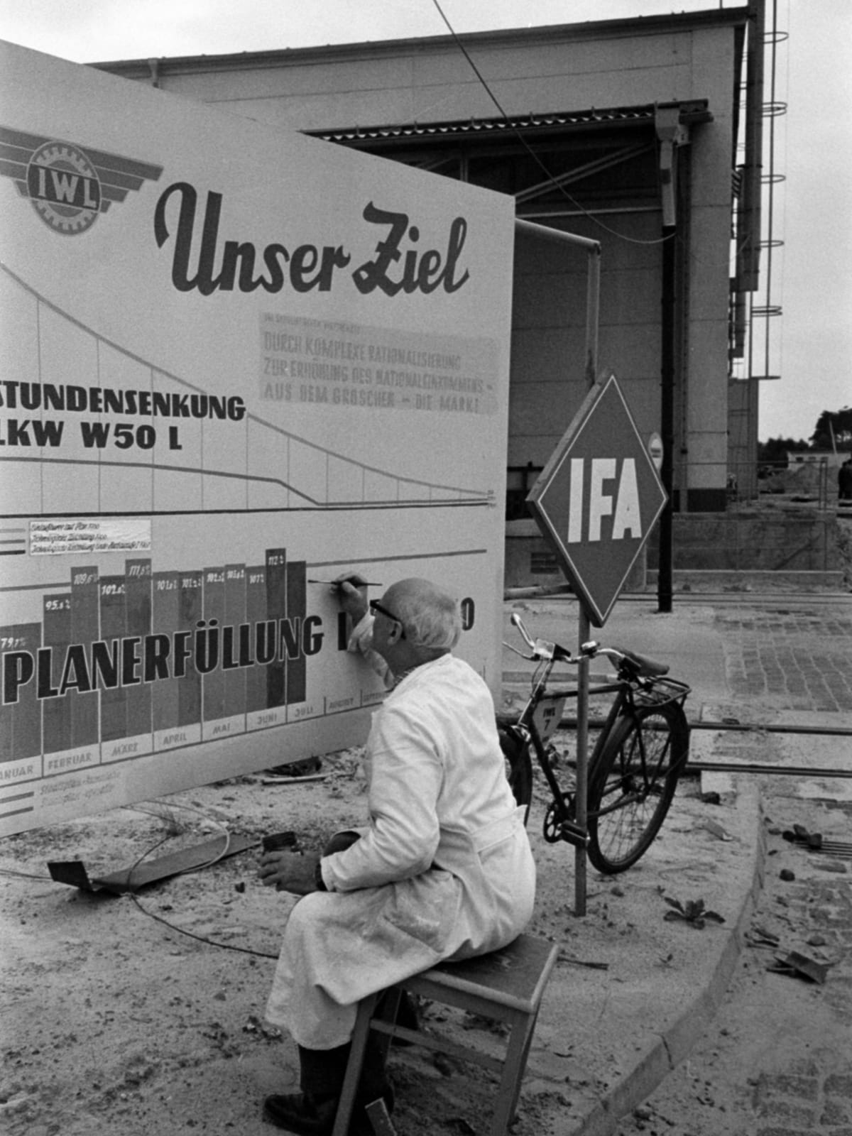 Black and white photo of a man on a stool painting a large standing sign board outside of an industrial building. There is a bicycle beside him, propped up on another freestanding sign with large letters that read IFA.