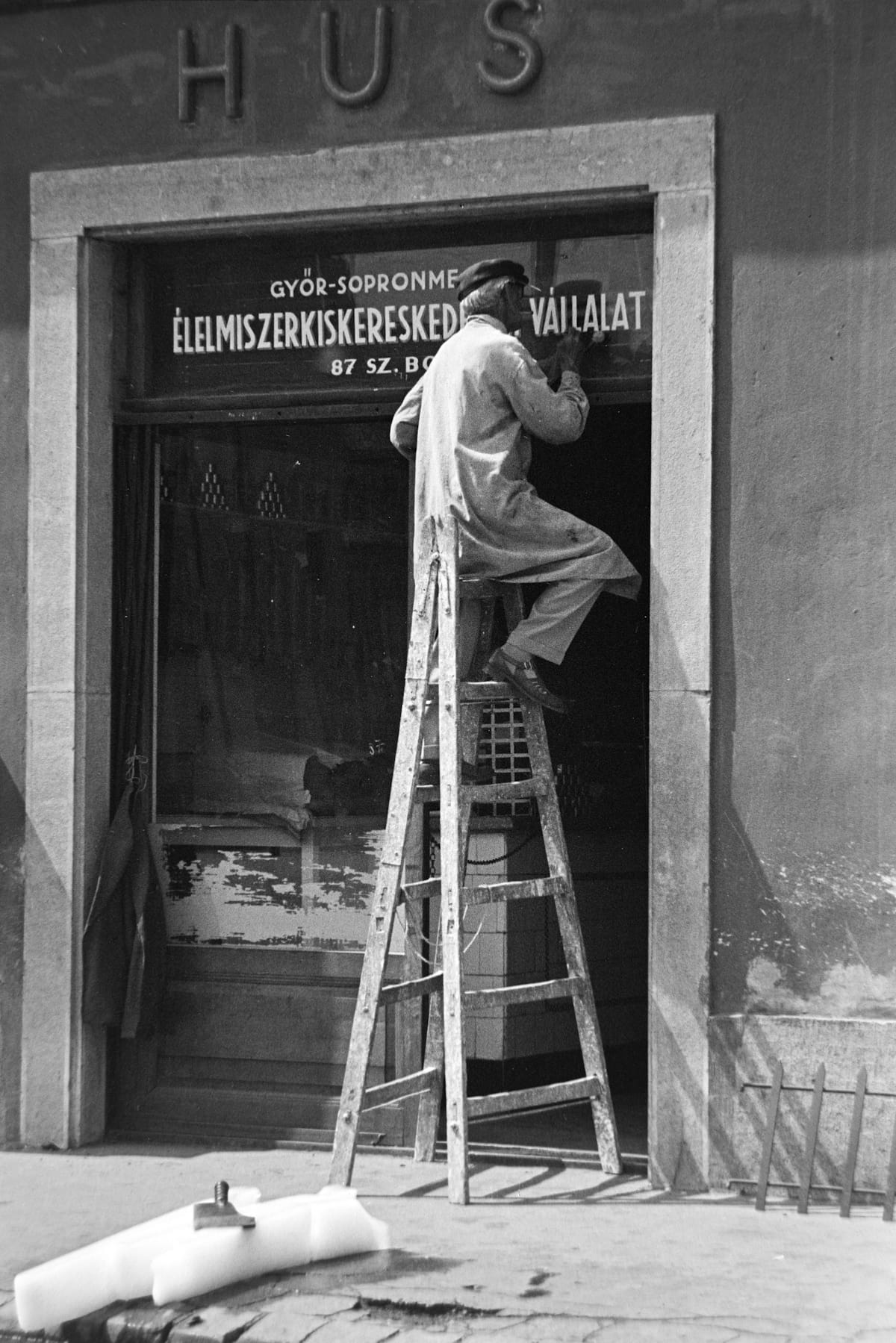 Black and white photo of a man straddling a ladder while painting the shadows onto lettering on a wide transom window.