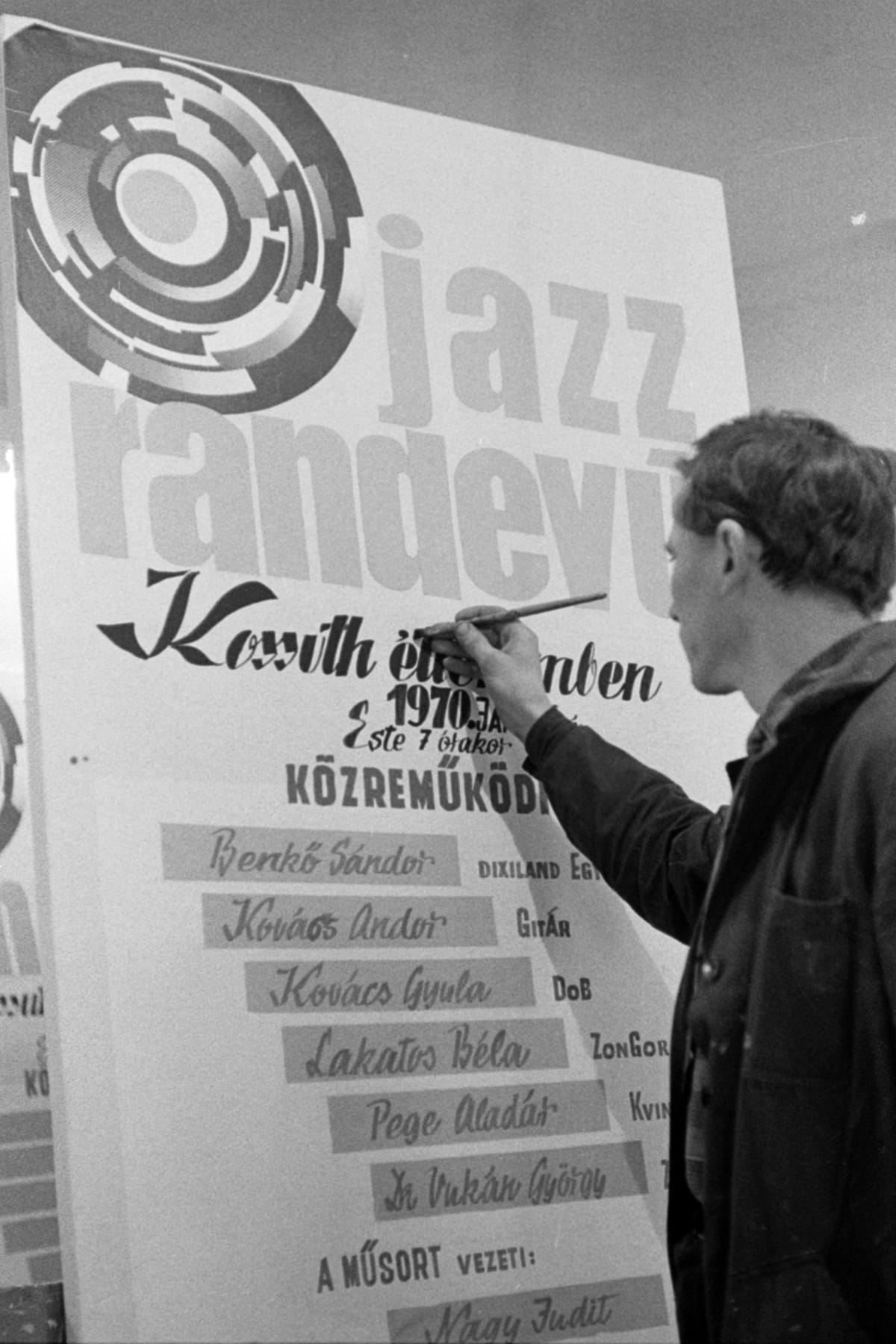 Black and white photo of a man adding the finishing touches to a lettered sign board advertising acts at a jazz festival.