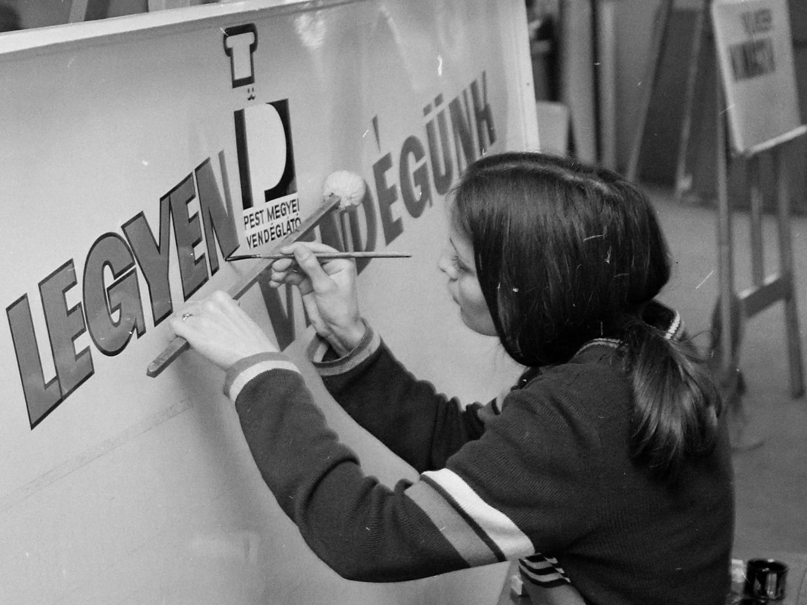 Over-the-shoulder view of a woman holding a mahl stick and painting lettering on a sign board.