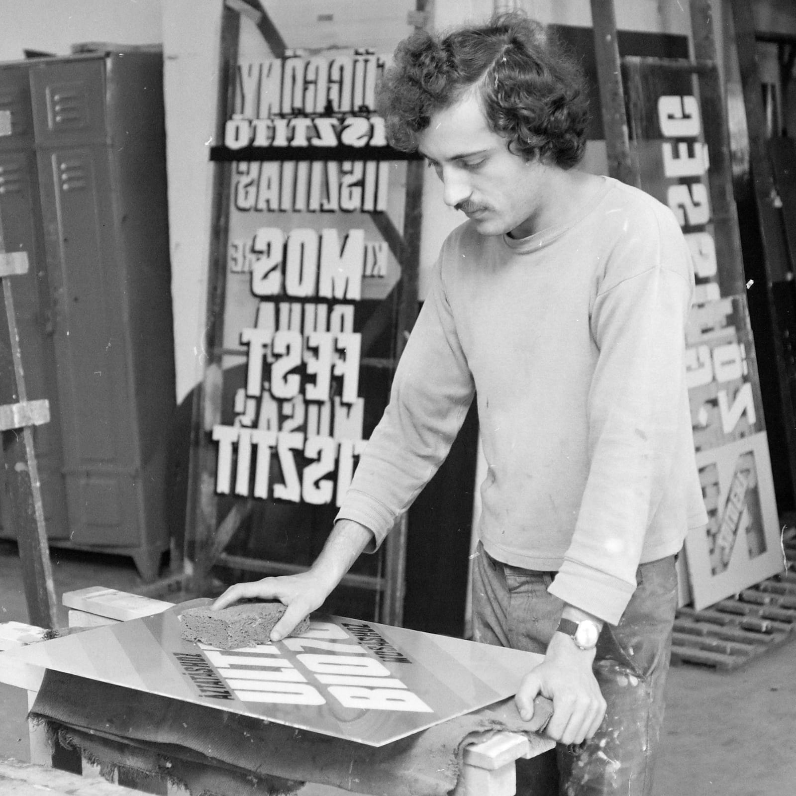 Black and white photo of a man stood over a workbench cleaning a lettered glass sign with a sponge. He is in a workshop setting and behind him are other signs and shop equipment.