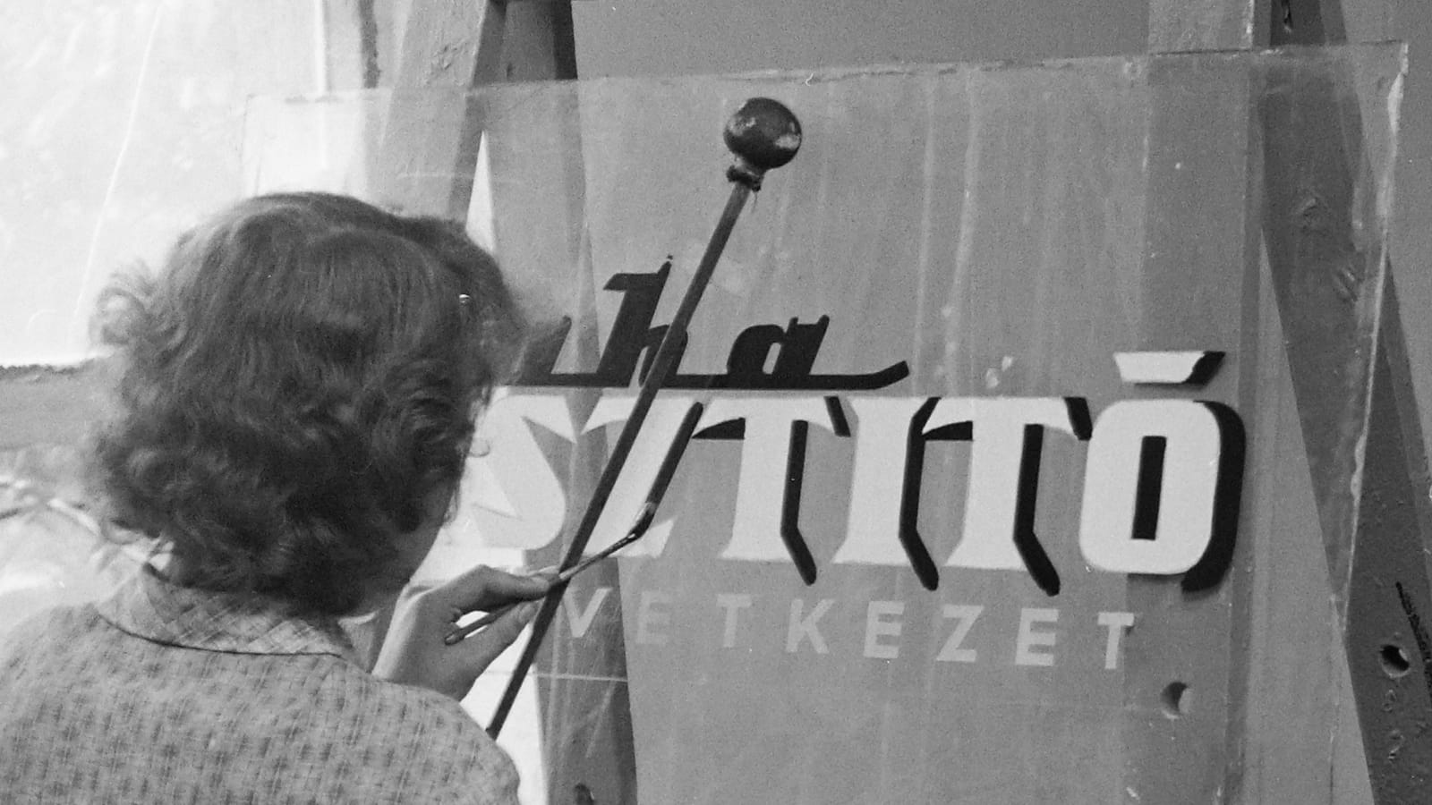 Black and white photo with a view from behind a woman adding dark shadows to lettering painted in white on glass.