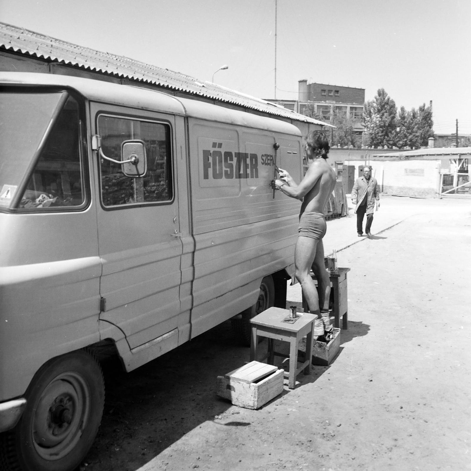 Black and white photo of a man wearing just boots and shorts while painting lettering onto the side of a van.