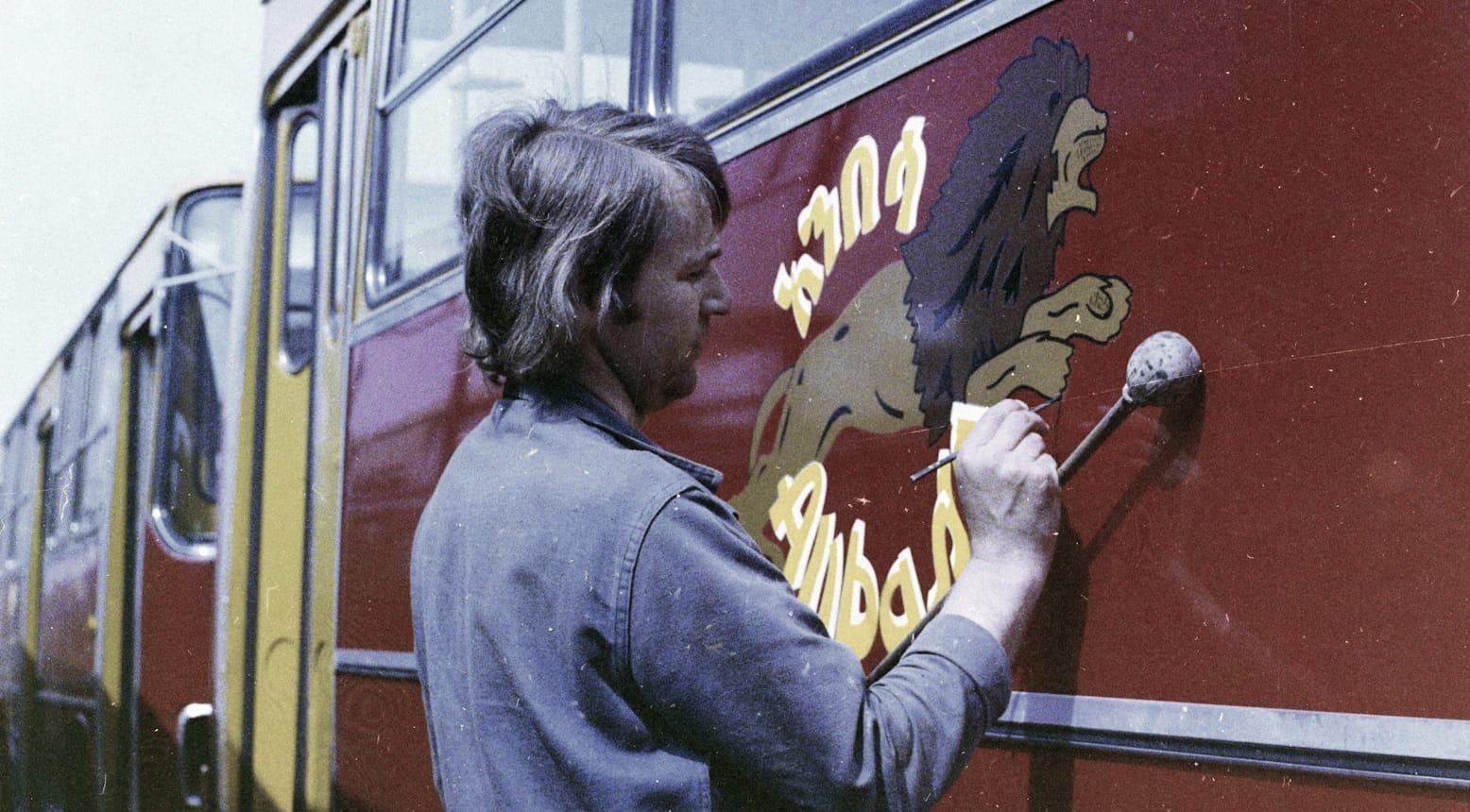 Grainy colour photo of a man painting a leaping lion on the side of a bus. Above and below these is lettering in yellow with a white shade.