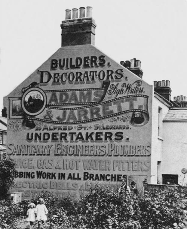 Black and white photo of a large gable-end wall adorned with painted lettering and pictorials advertising the many services of the Adams & Jarrett firm. These include building, decorating, signwriting, undertaking, plumbing, sanitary engineering, and more.