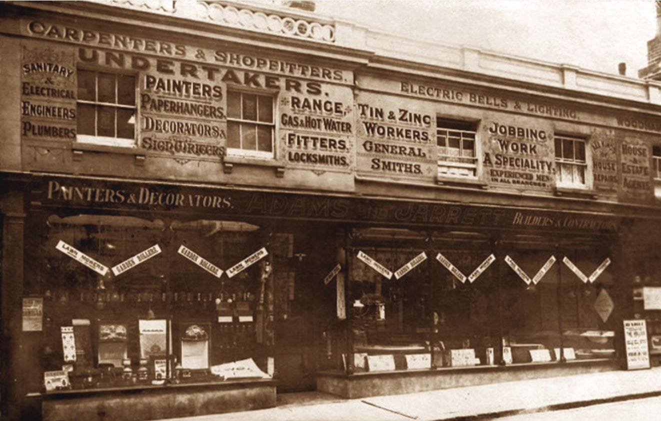 Black and white photo of the Adams & Jarrett shop. It's set across the width of two buildings, and above their fascia sign are panels of decorative lettering between first floor windows. These advertise their various services across a variety of decorative painting and building trades.