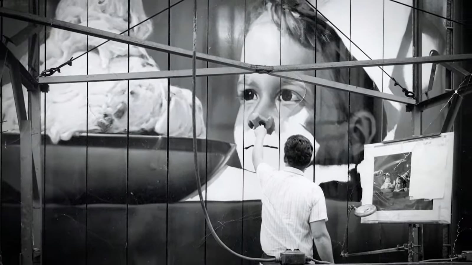 Black and white photo showing the view from behind a man painting a boy's face looking at a bowl of what looks like ice cream.