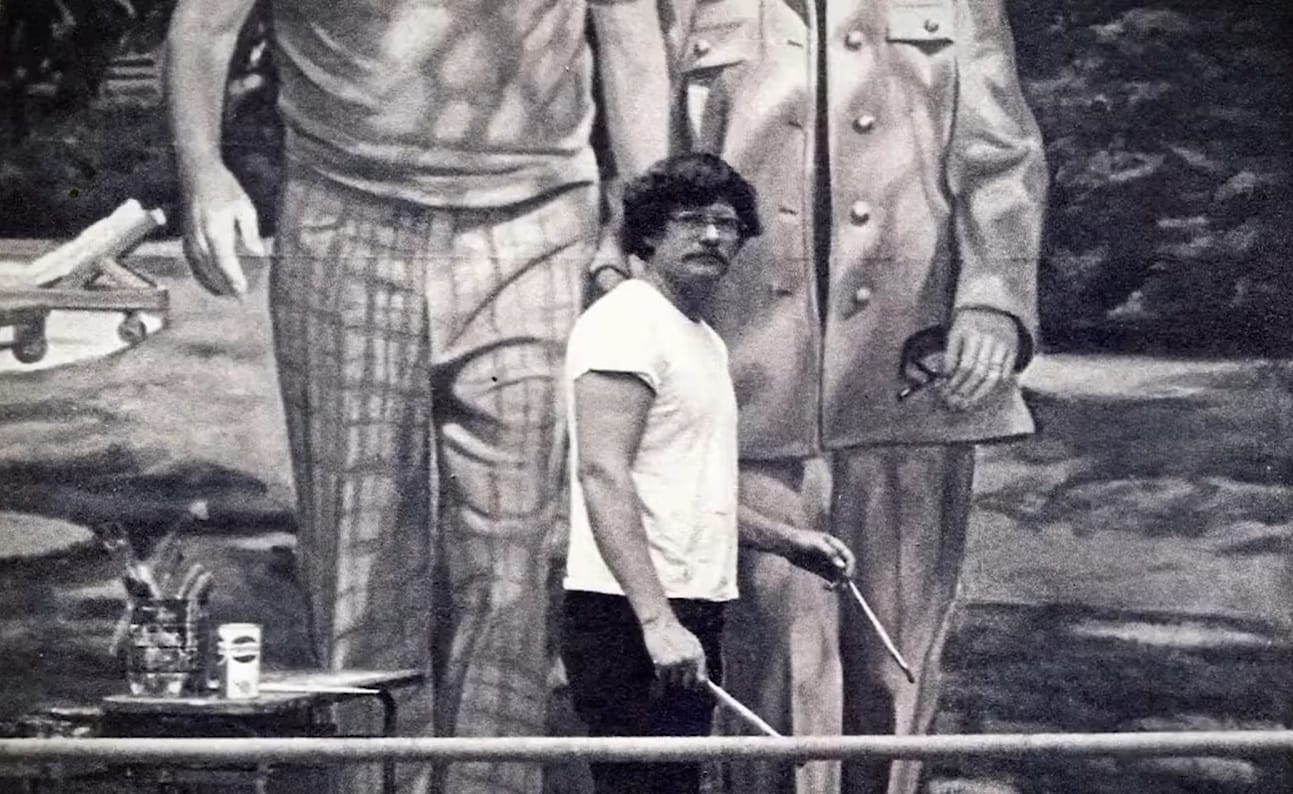 Black and white photo of an artist posing with his brushes in front of a large-scale painting of two people walking side by side.