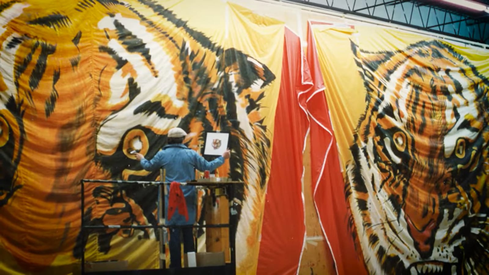 Colour photo of a man on a platform painting one of two close-ups of a tiger's face on fabric.