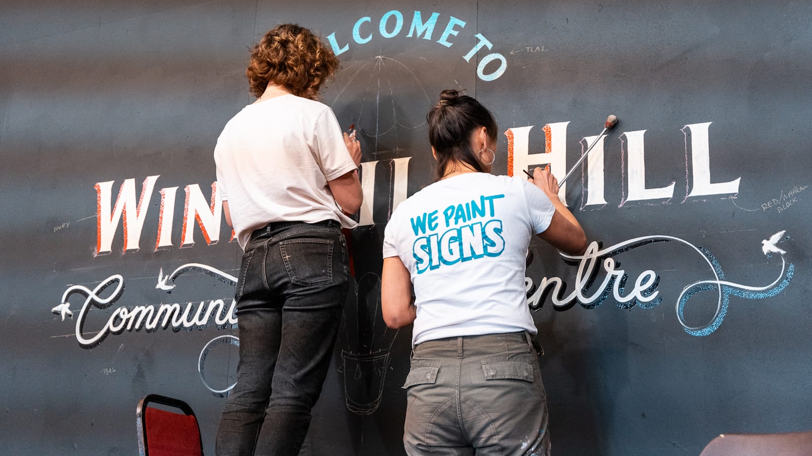 View from behind two women painting a mural on a black wall. The mural says "Welcome to Windmill Hill Community Centre".