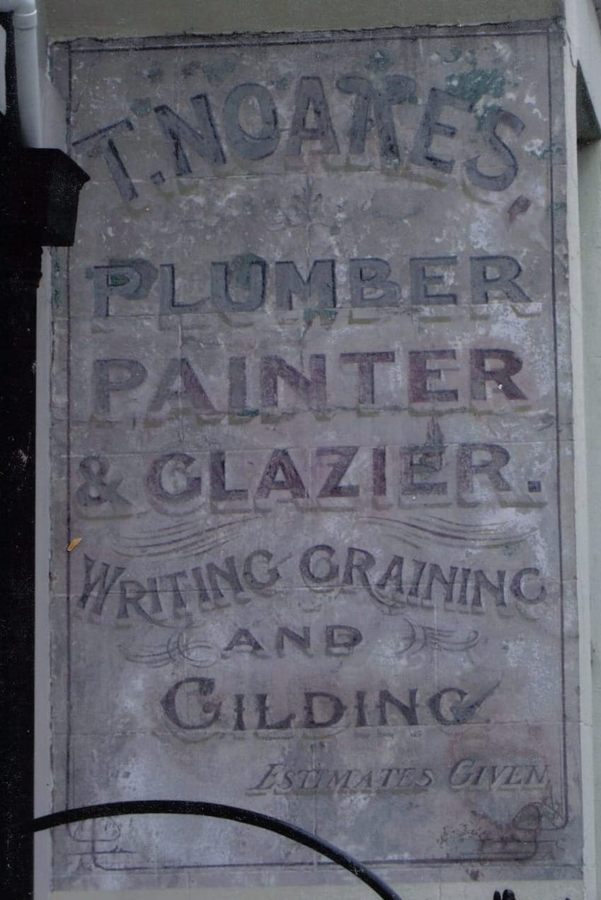 Fading painted sign on a stucco wall with decorative lettering that reads: "T. Noakes, Plumber, Painter, & Glazier, Writing, Graining, and Gilding, Estimates Given."