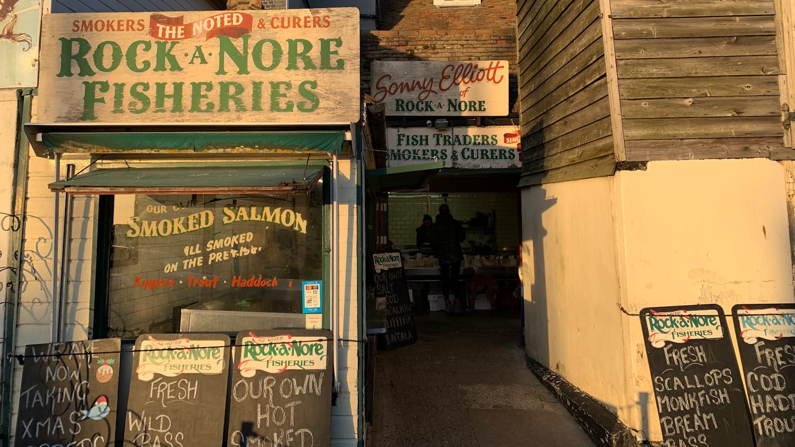 Various bits of hand-painted signs advertising the Rock-a-Nore Fisheries, including their fascia sign, painted window, blackboards lined up on the street, and then signs above the entrance which is set back from the main building frontage.