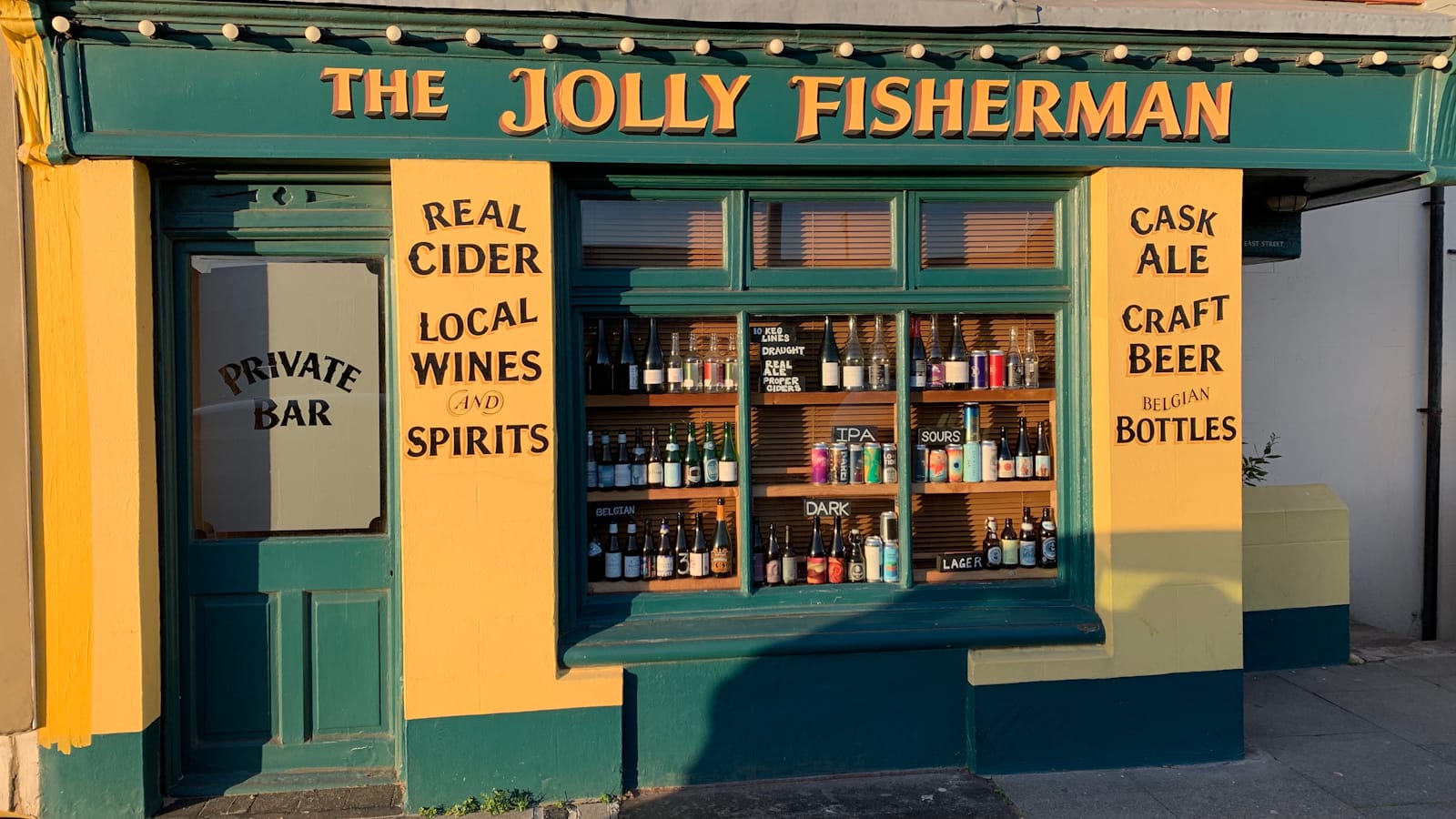 Bar frontage with a fascia sign, The Jolly Fisherman, and panels of lettering on the pillars that site either side of the main central window. The window has various bottles of beer on display on shelves, while the pillar lettering reads "Read Cider, Local Wines and Spirits" (left) and "Cask Ale, Craft Beer, Belgian Bottles" (right).