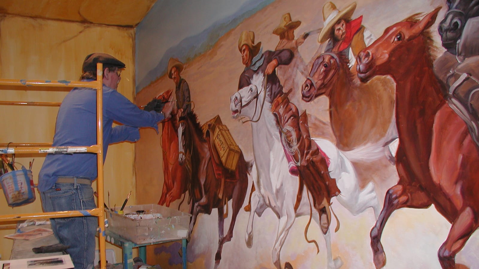 Man painting a mural showing cowboys galloping on horses.