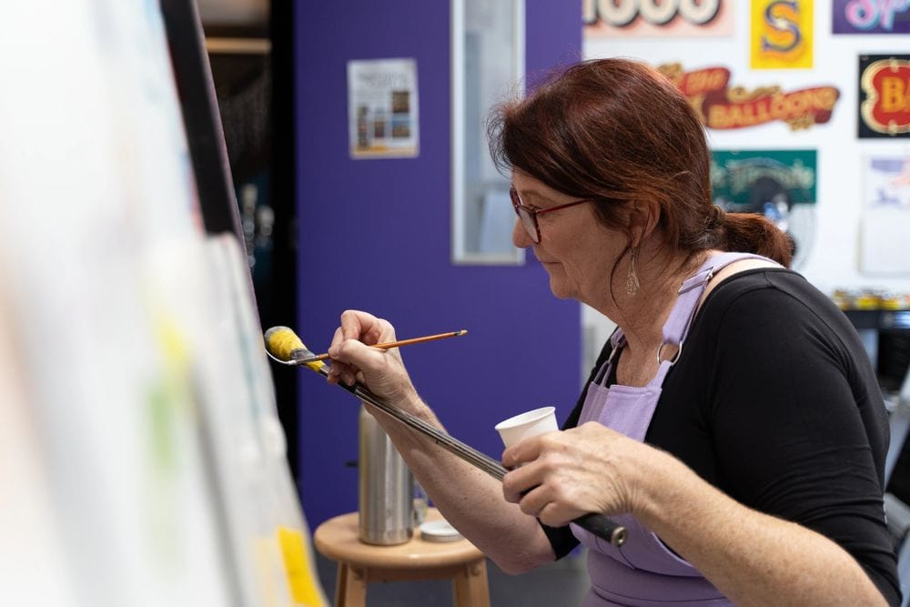 Profile of a woman using a mahl stick to steady her hand as she paints lettering onto a board.