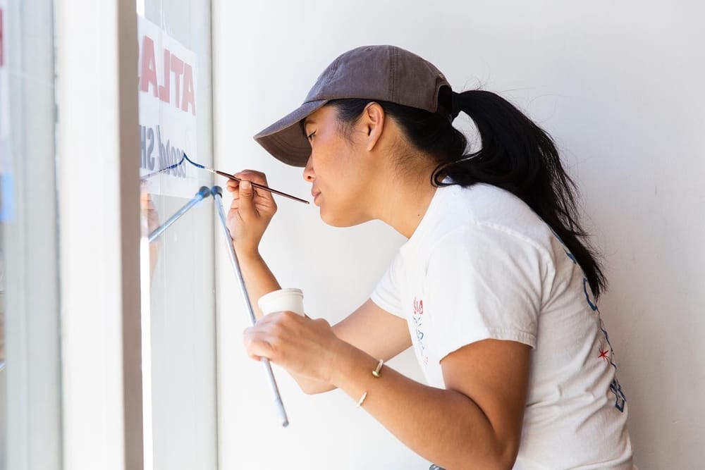 Woman in a baseball cap focused on painting blue lettering in reverse on a window.